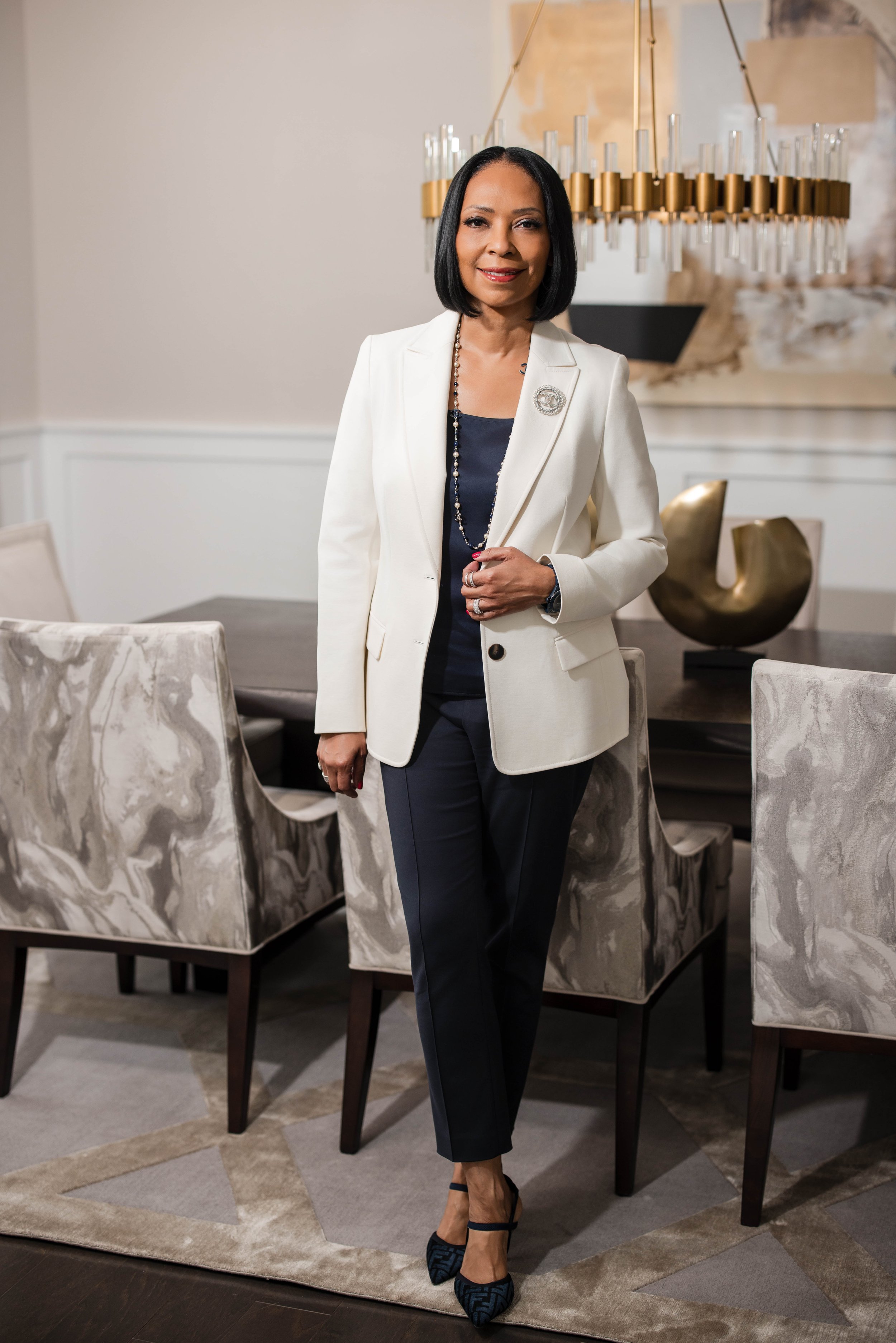 A woman with short black hair, wearing a white blazer with a decorative brooch, a black top, black pants, and high-heeled shoes standing in a dining room with a chandelier, a large abstract painting, and patterned chairs.