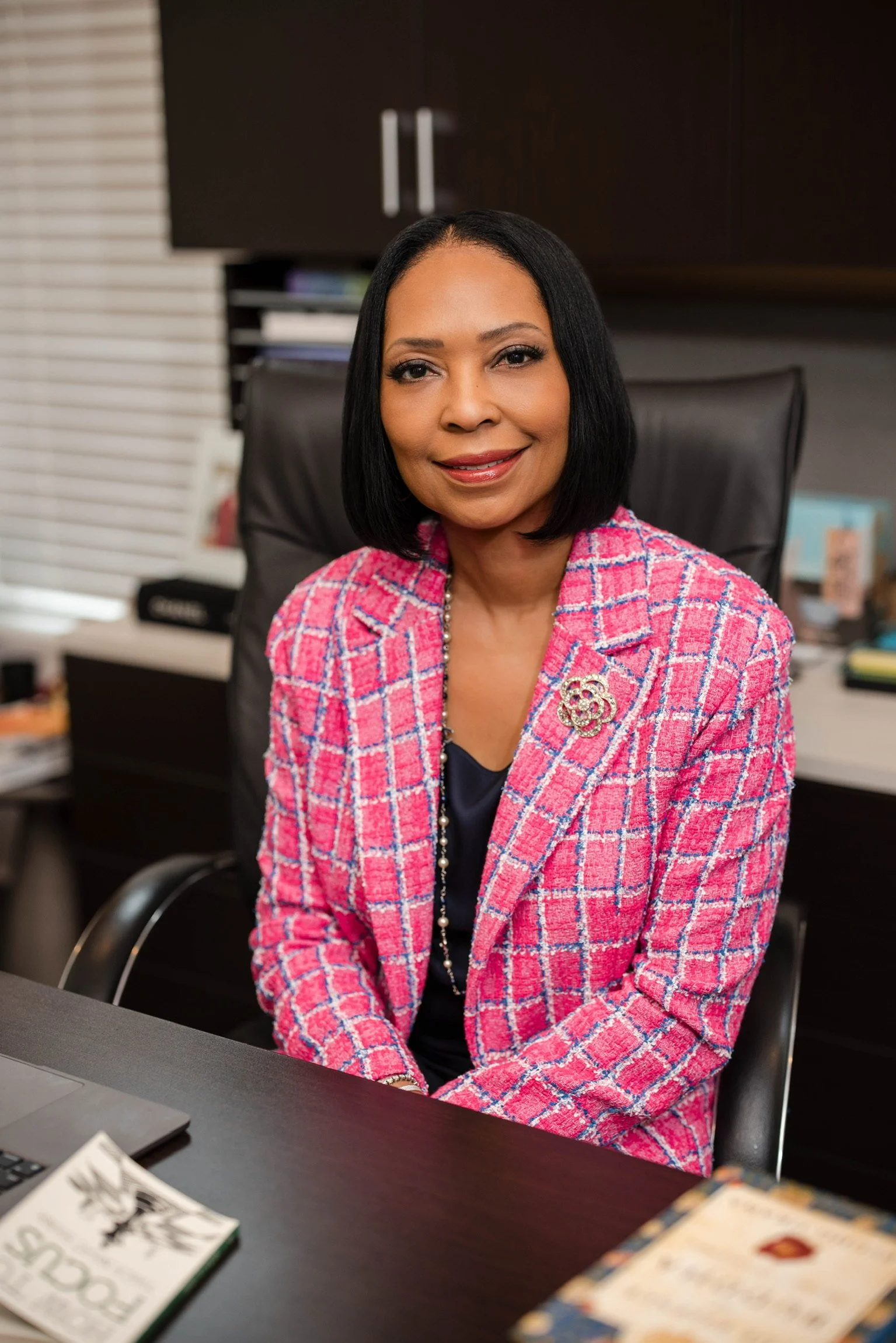 A woman with medium skin tone and black hair, wearing a pink and red plaid blazer with a brooch, sitting at a desk in an office with a black chair and dark cabinets in the background.