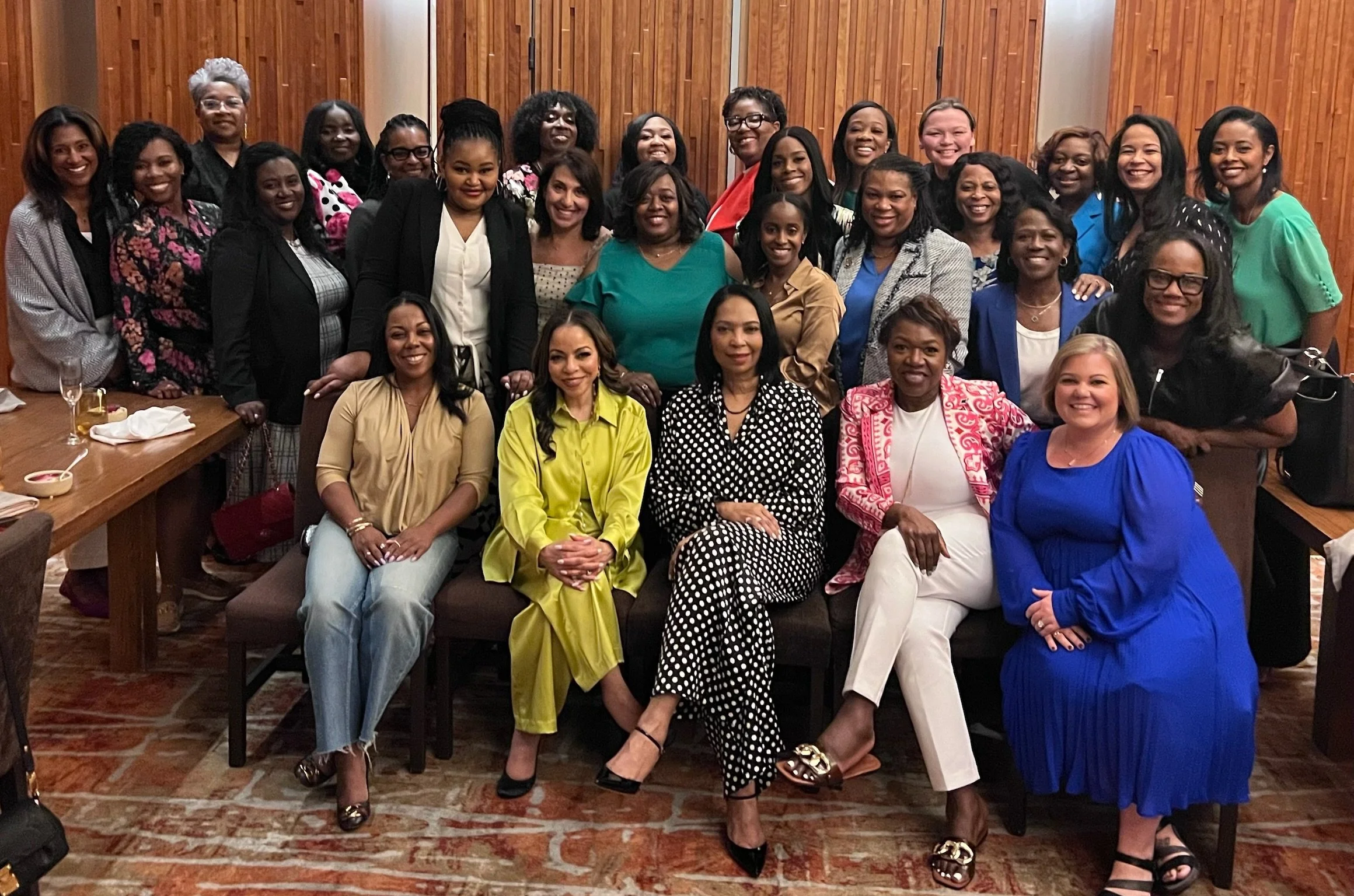 A large group of women gathered together for a photo indoors, smiling and dressed in colorful and professional attire, with wooden wall paneling in the background.