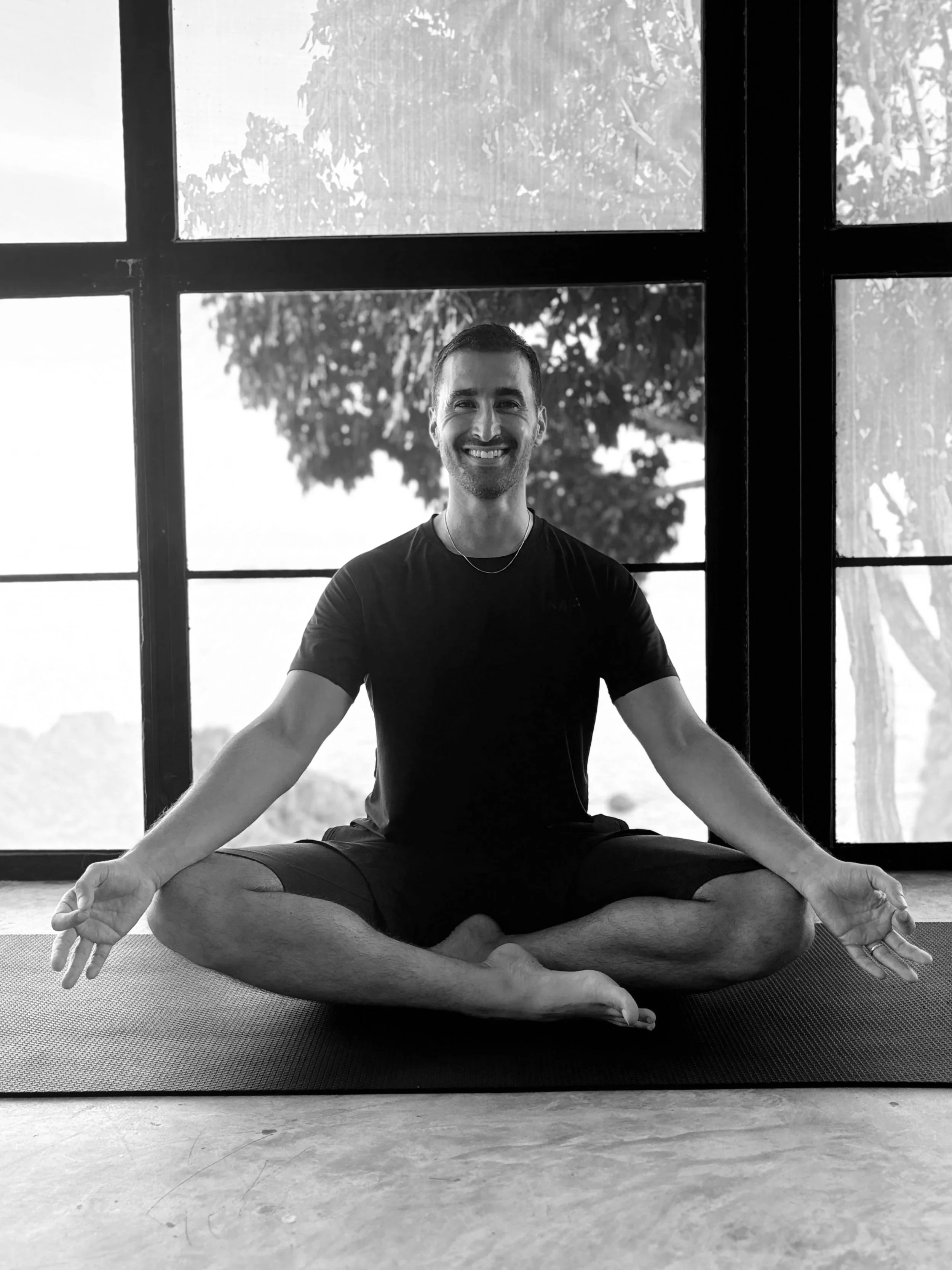 Dr. Moaeri macht Yoga im Sitzen im Schneidersitz auf einer Matte vor großen Fenstern, lächelnd, in einem Raum mit natürlichem Licht.