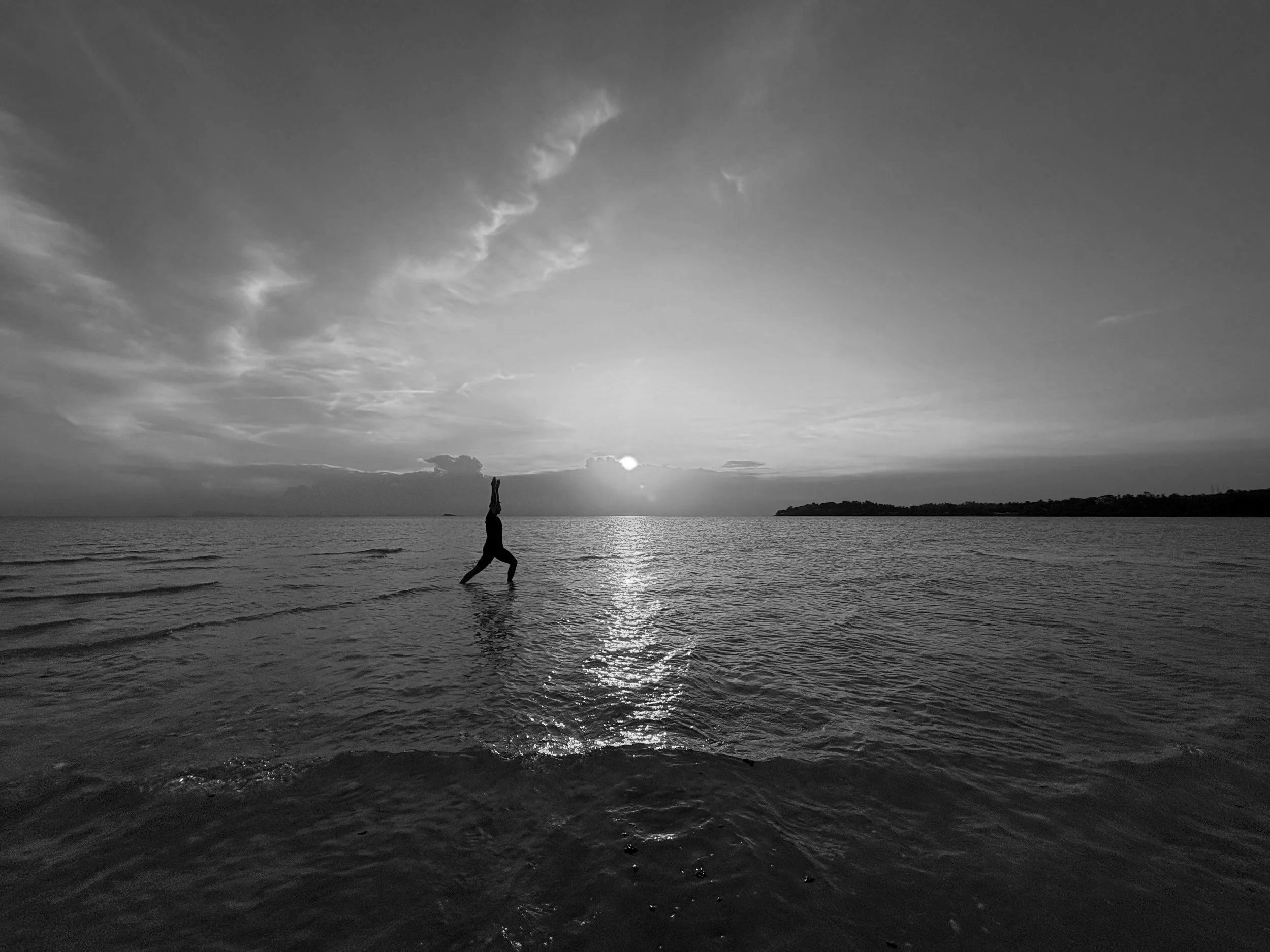 Dr. Moaeri  steht in flachem Wasser am Strand und macht Yoga, während die Sonne untergeht oder aufgeht, im Hintergrund ein Horizont mit Wolken und einem Landstreifen.