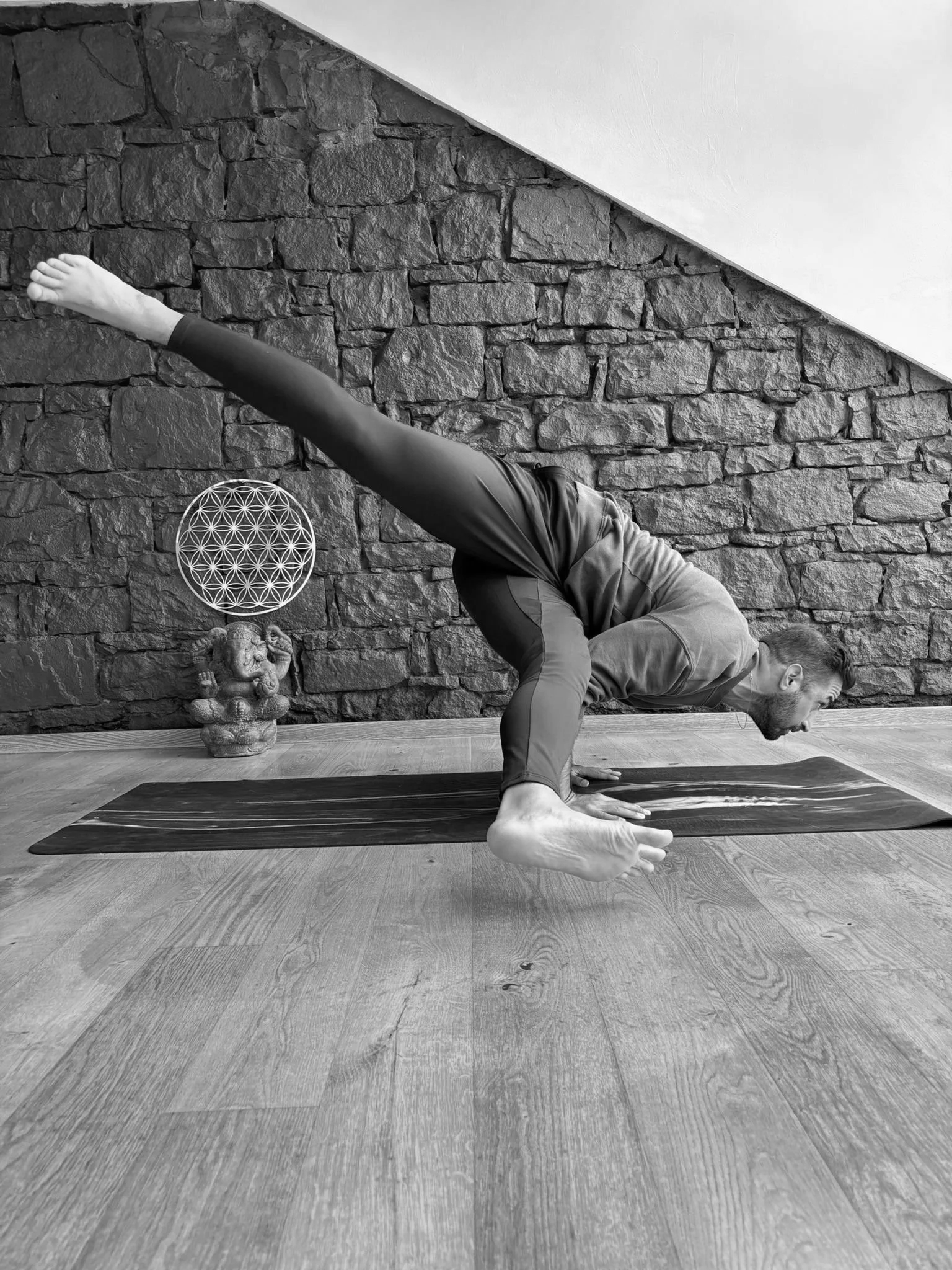 Dr. Moaeri macht Yoga mit Arm-Balance Übung auf einer Yoga-Matte in einem Raum mit Steinwand und Holzfußboden.