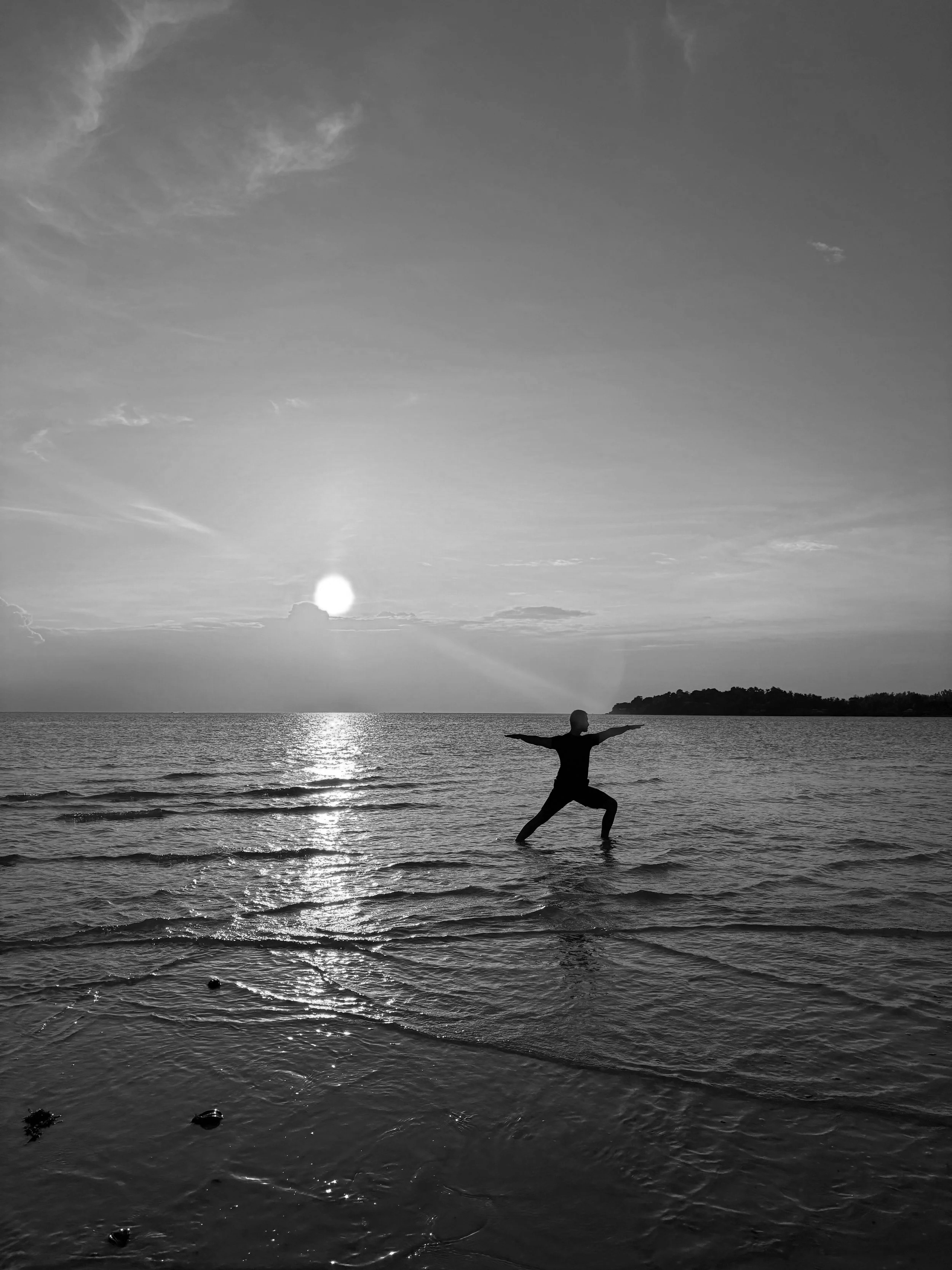 Ein Mensch steht im Wasser am Strand, hält die Arme breit, beim Sonnenuntergang am Meer, schwarz-weiße Fotografie.