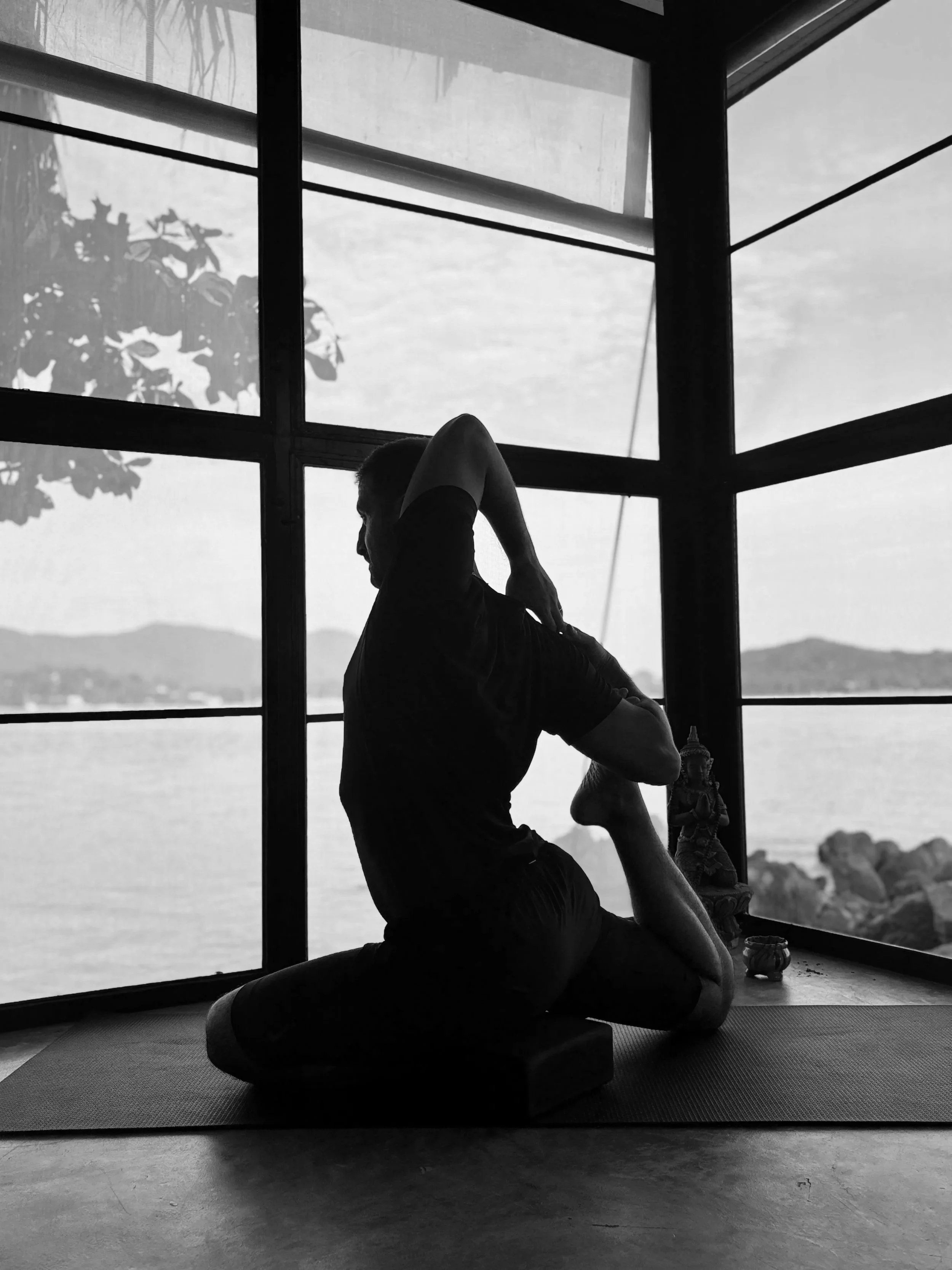 Mensch übt Yoga in einem Raum mit großen Fenstern und Blick auf Wasser und Berge, bei Sonnenlicht.