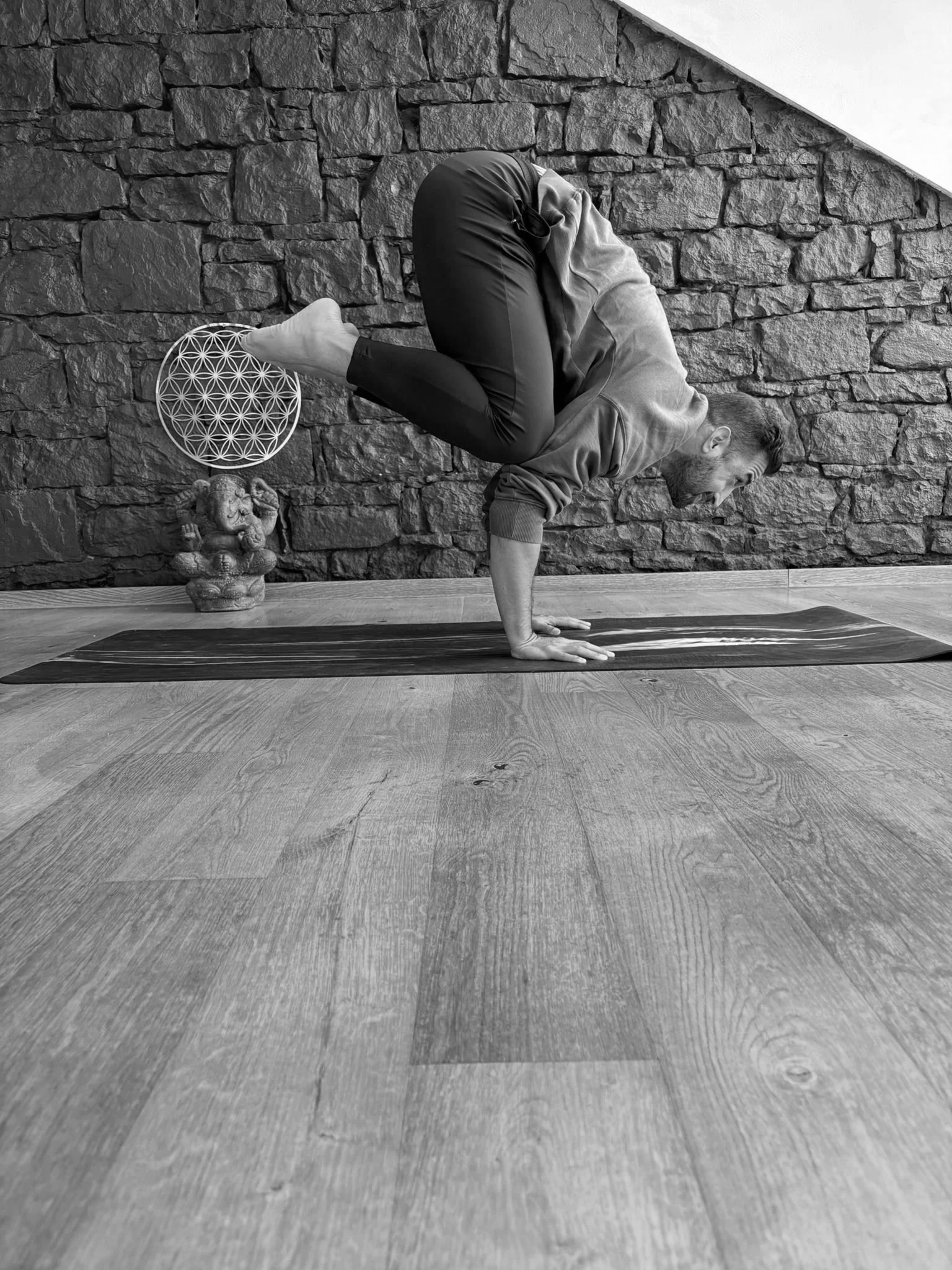 Ein Mann macht einen Handstand auf einer Yoga-Matte vor einer Steinwand, mit einer kleinen Statue und einer geometrischen Dekoration im Hintergrund.