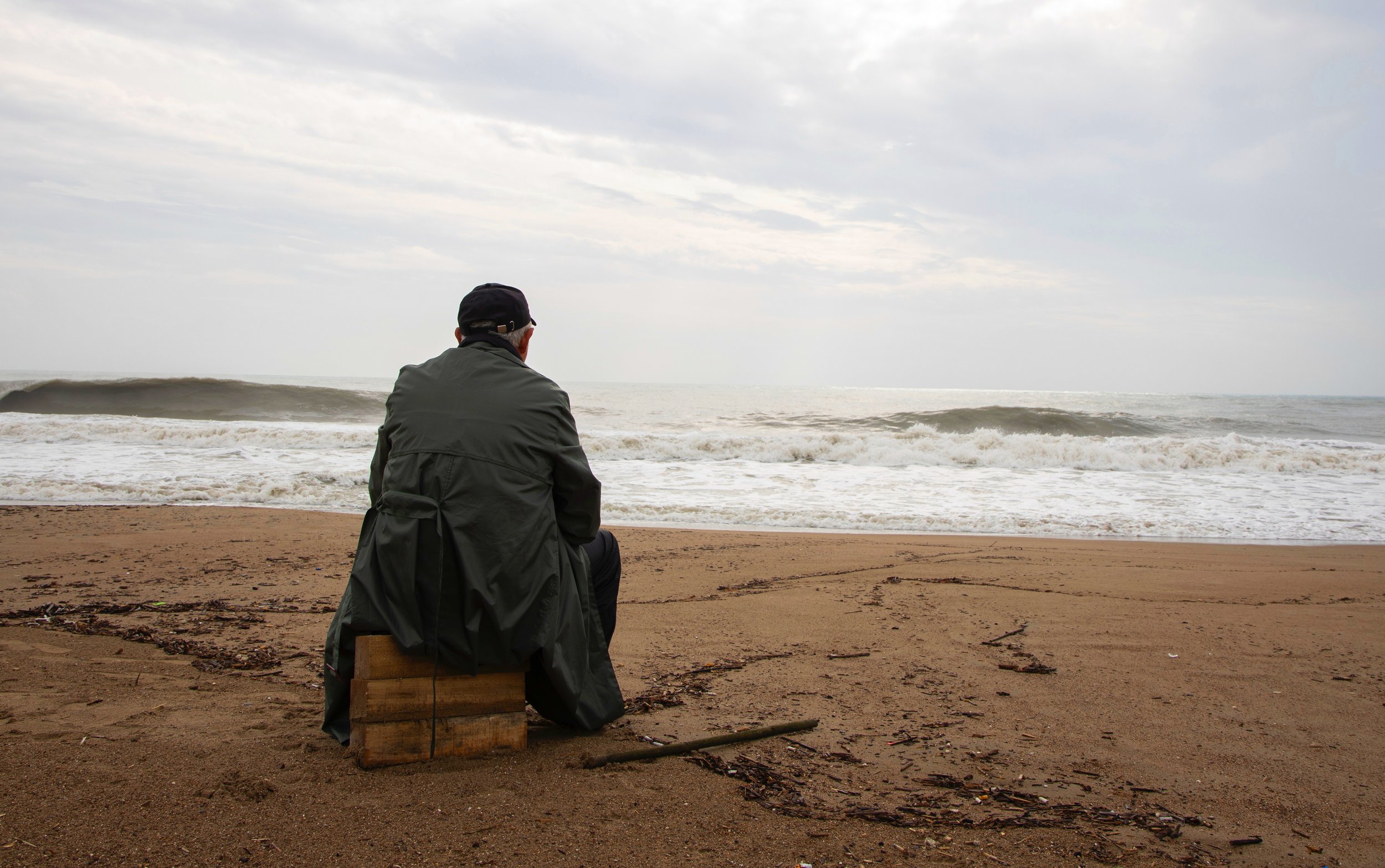 An elderly man wearing a dark jacket and cap sitting on a wooden block on a sandy beach, looking at ocean waves under a cloudy sky.