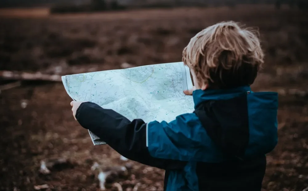 A young boy with blond hair, wearing a blue and black jacket, is standing outdoors and holding a map, looking at it.