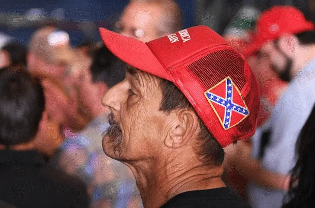 A man wearing a red cap with a Confederate flag patch, surrounded by other people at a gathering or event.