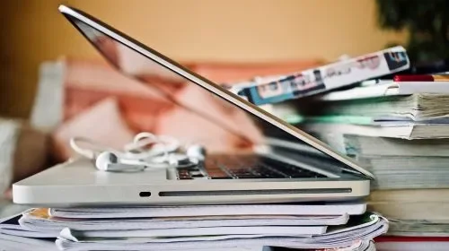 A laptop placed on top of stacks of papers and books on a desk.