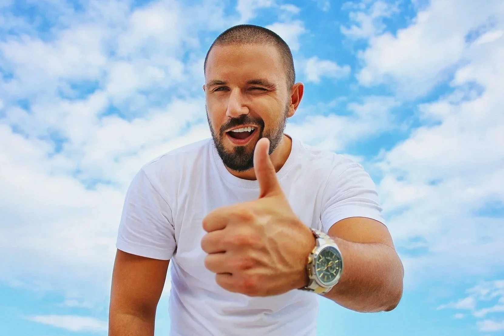 A man with a beard and short hair, wearing a white T-shirt and a wristwatch, showing a thumbs-up gesture with his right hand, smiling and winking against a blue sky with scattered white clouds.