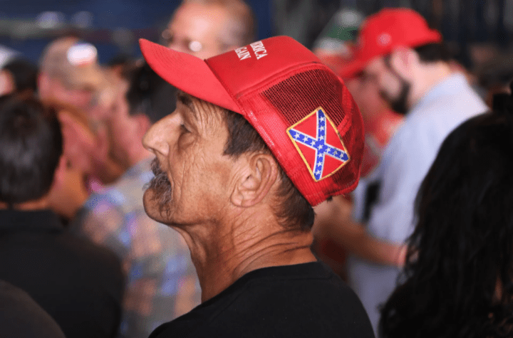 A man wearing a red baseball cap featuring the Confederate flag, surrounded by other people at a gathering or event.