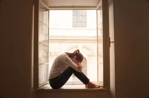 A person sitting alone on an open windowsill with knees drawn to chest, indoors during daytime.