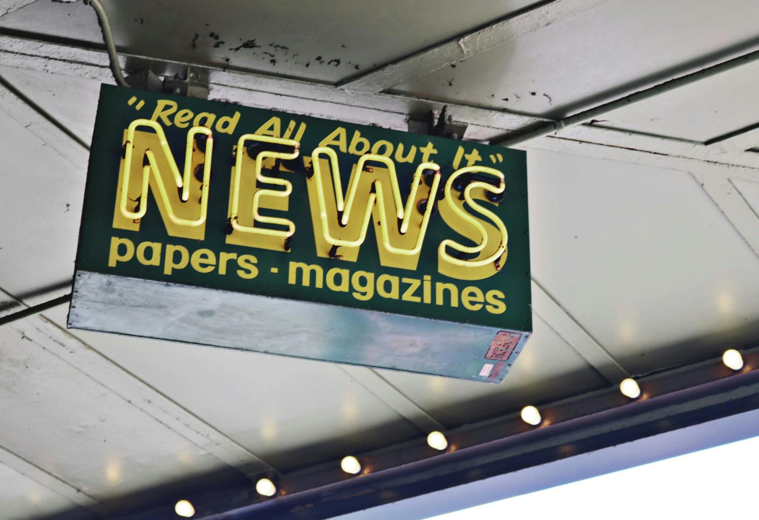 A street sign with yellow neon lights spelling 'NEWS' and the words 'Read All About It' and 'papers - magazines' written on a green background.