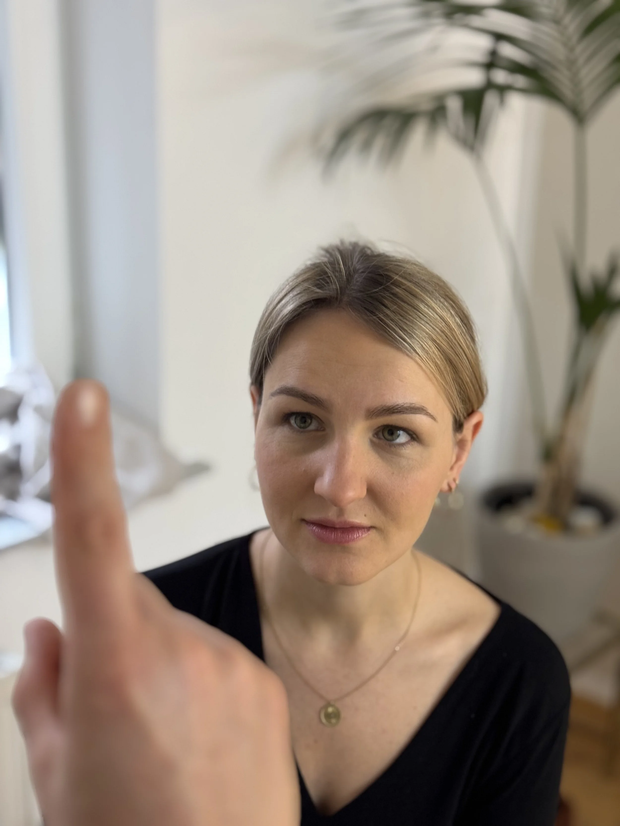 Eine Frau mit lockerer, heller Haarfarbe und dunklem Top, die vor einem Raum steht, blickt aufmerksam und freundlich, während jemand anderes im Bild eine Hand mit einem Tuch oder Papier hochhält.