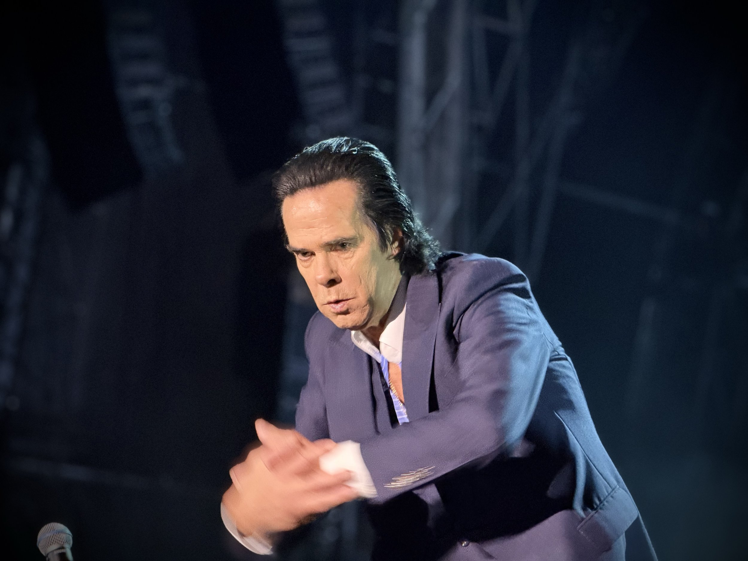 A man with dark, slicked-back hair wearing a purple jacket and white shirt, looking down while sitting at a desk with a microphone nearby.