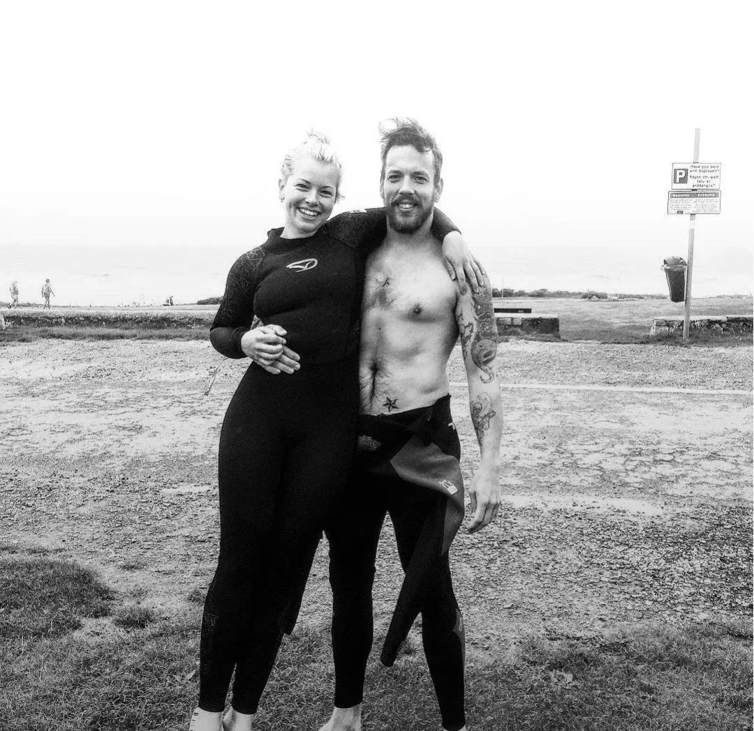 A smiling woman in a wetsuit and a shirtless man with tattoos on a beach, standing close together with their arms around each other.