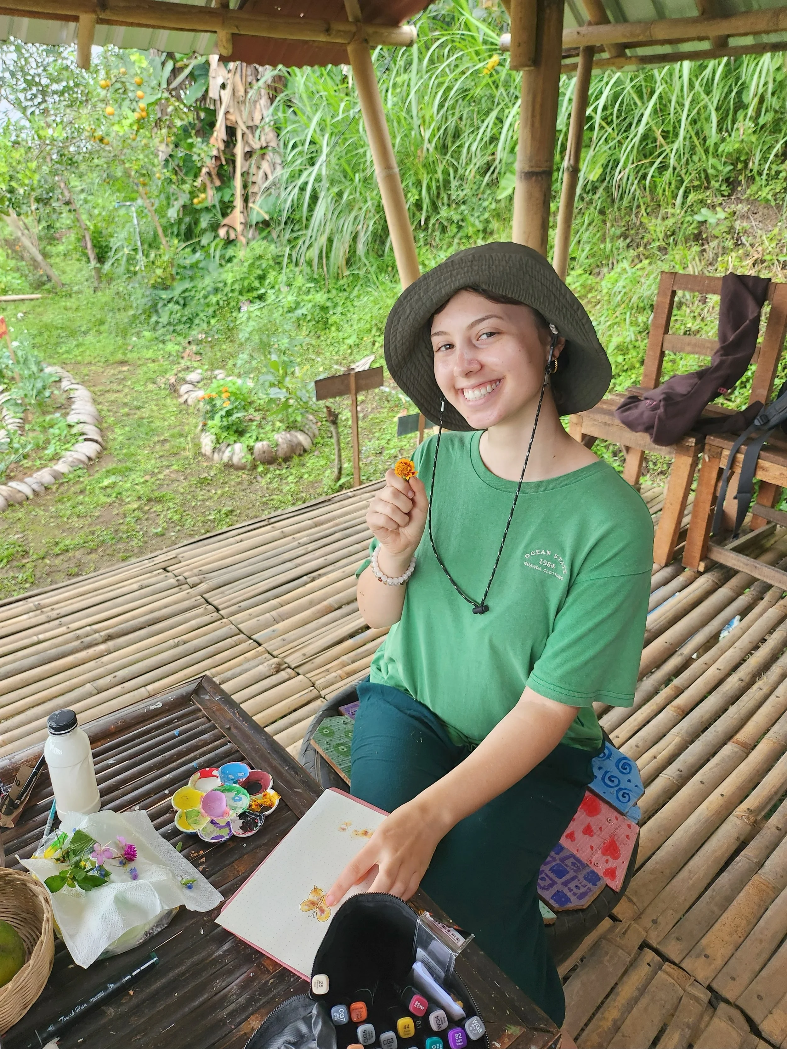 A young woman with short hair smiling and holding a small flower, sitting at a bamboo table outdoors with art supplies nearby, surrounded by lush green plants and a wooden structure.