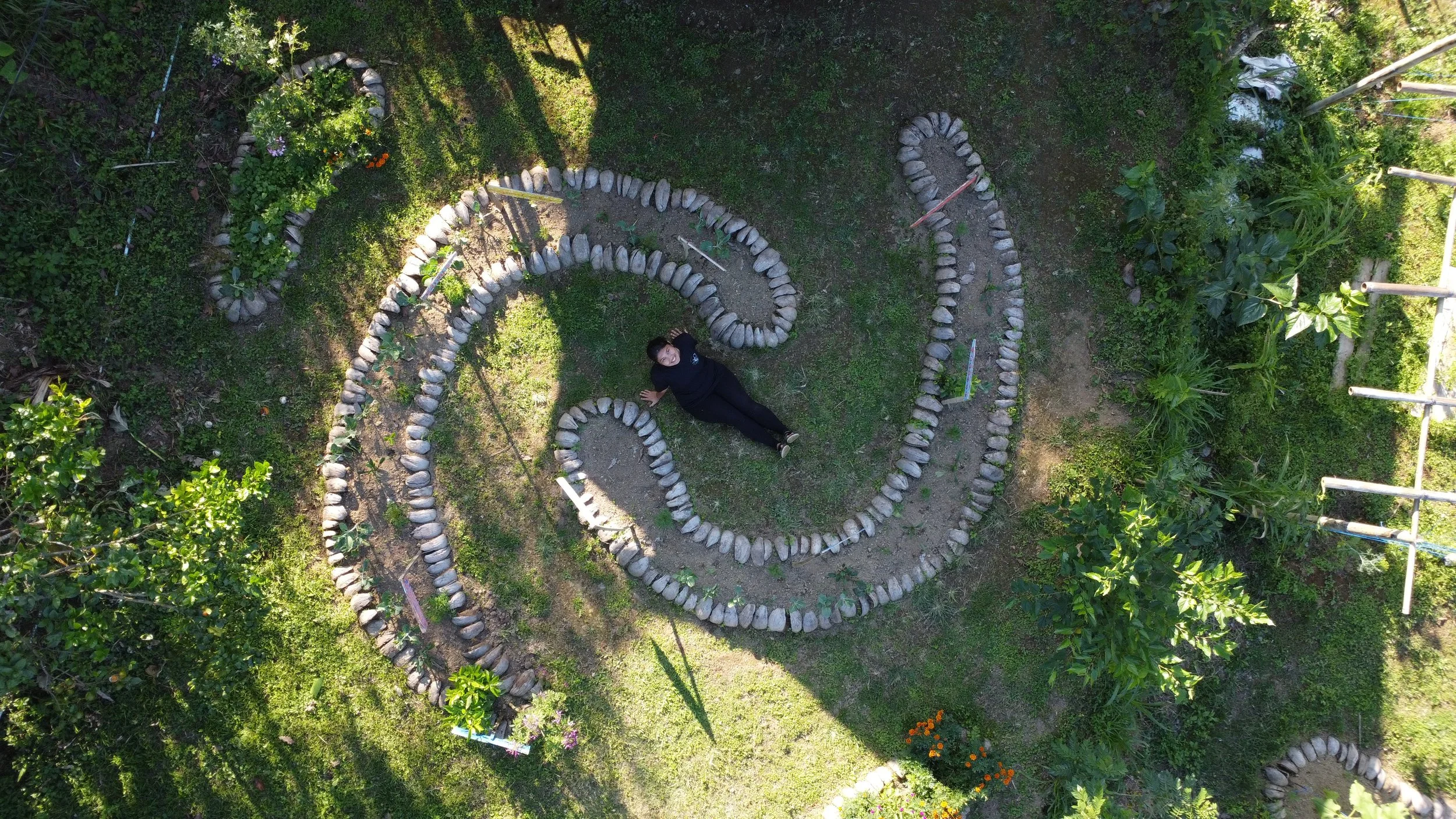 A woman is lying down in a garden, surrounded by a labyrinth formed from rocks. The garden is lush with greenery and flowers, and the labyrinth has a spiraling pattern.