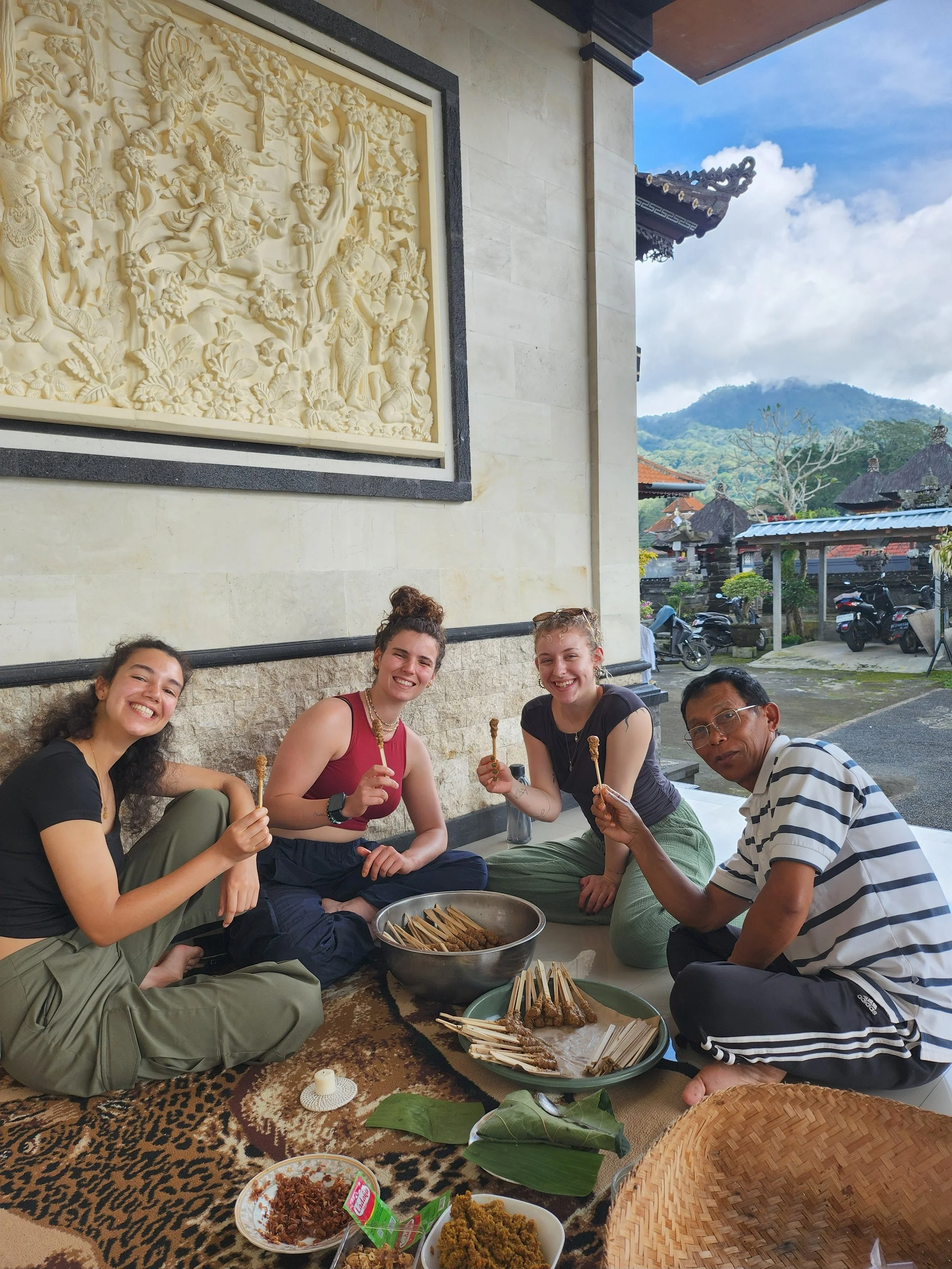 Four smiling people sitting on the floor with traditional Indonesian food, skewers, and dishes in front of them, indoors with a mountain view outside.