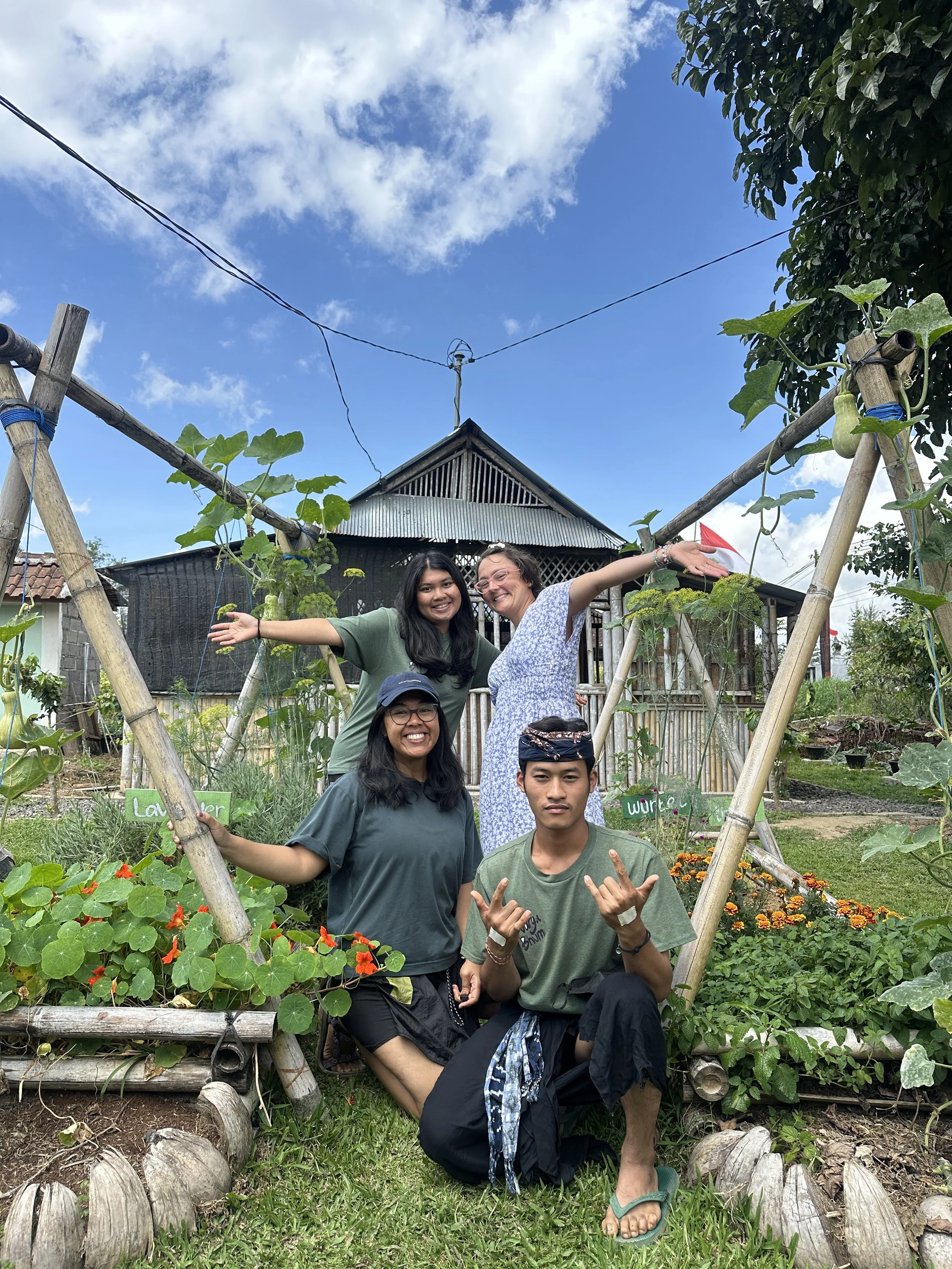 Four people smiling in a garden with a bamboo arch and a rustic wooden house in the background.