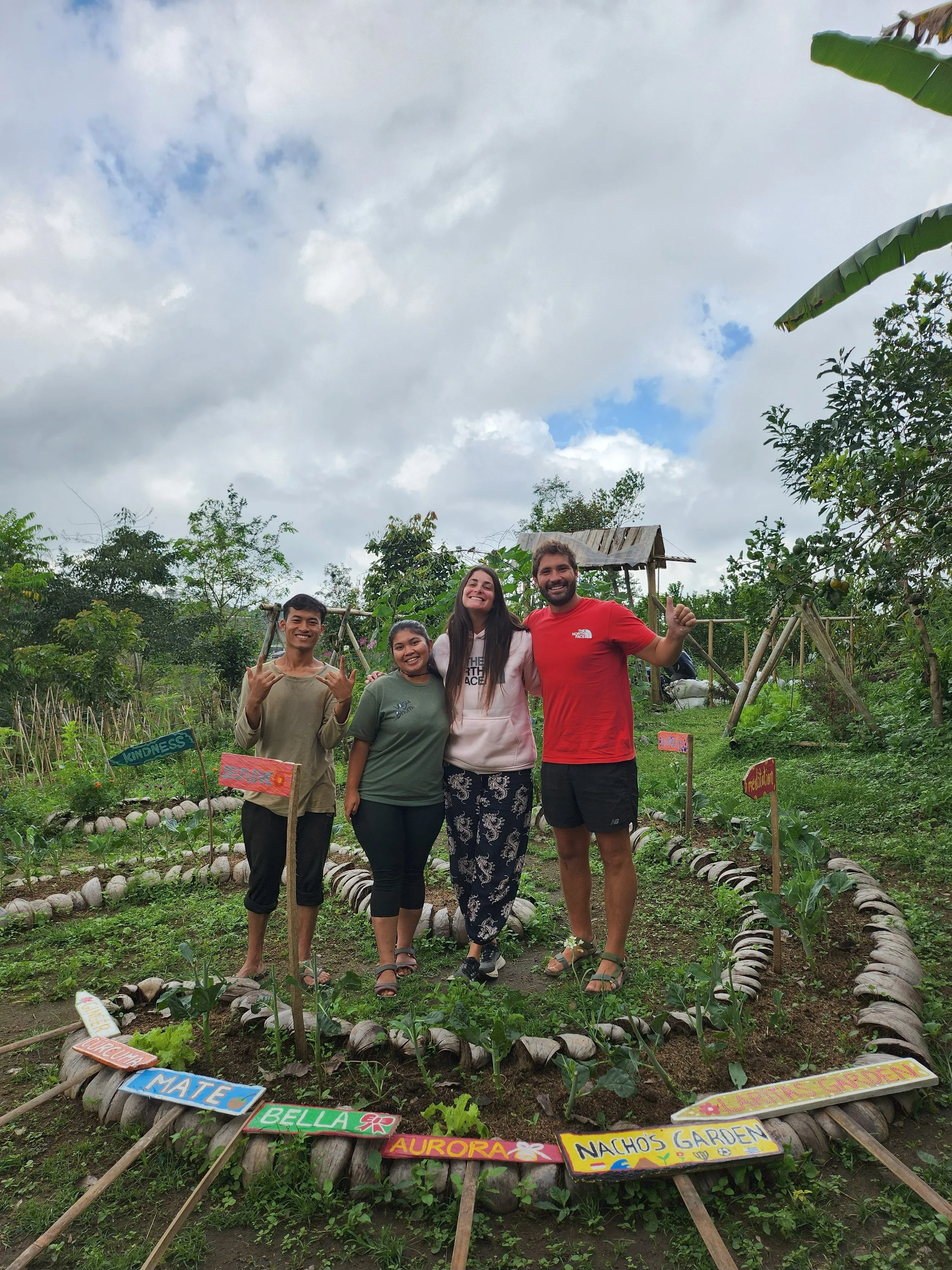 Four people standing together in a community garden, smiling and posing for the photo. The garden has various plants and colorful signs with names such as 'MATE', 'BELLA', 'AURORA', and 'NACHO'S GARDEN'.