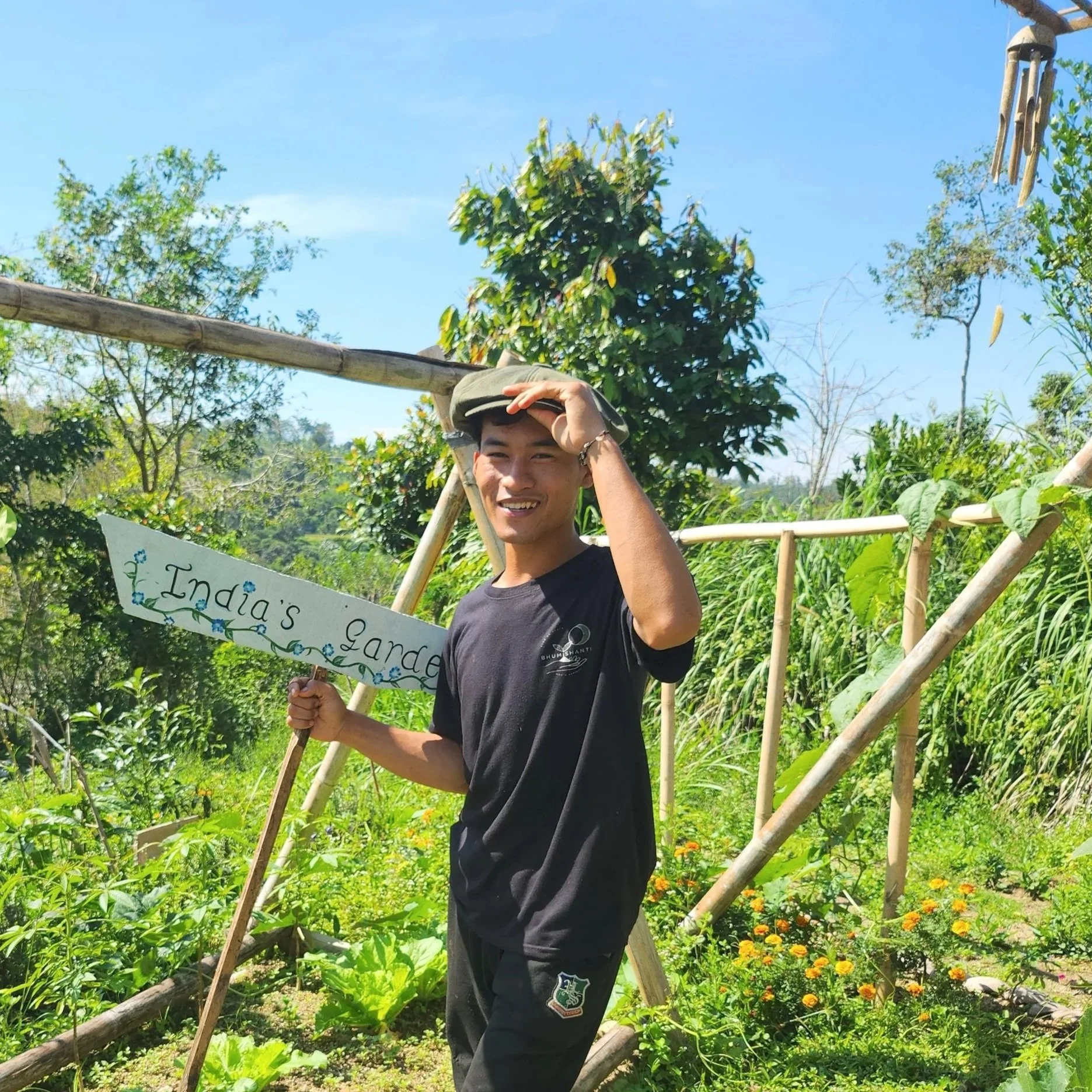 A young man standing in a lush garden holds a sign that reads 'India's garden' and smiles at the camera. He wears a black t-shirt and a cap, with greenery and wooden structures in the background under a blue sky.