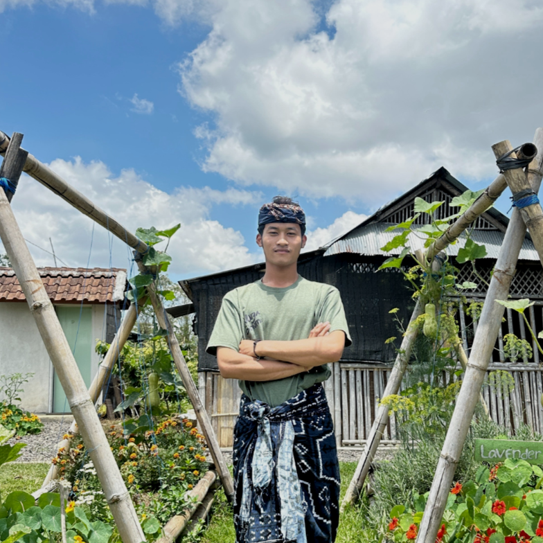 A young man standing in a garden with wooden trellises for plants, surrounded by flowers and greenery, under a partly cloudy sky.