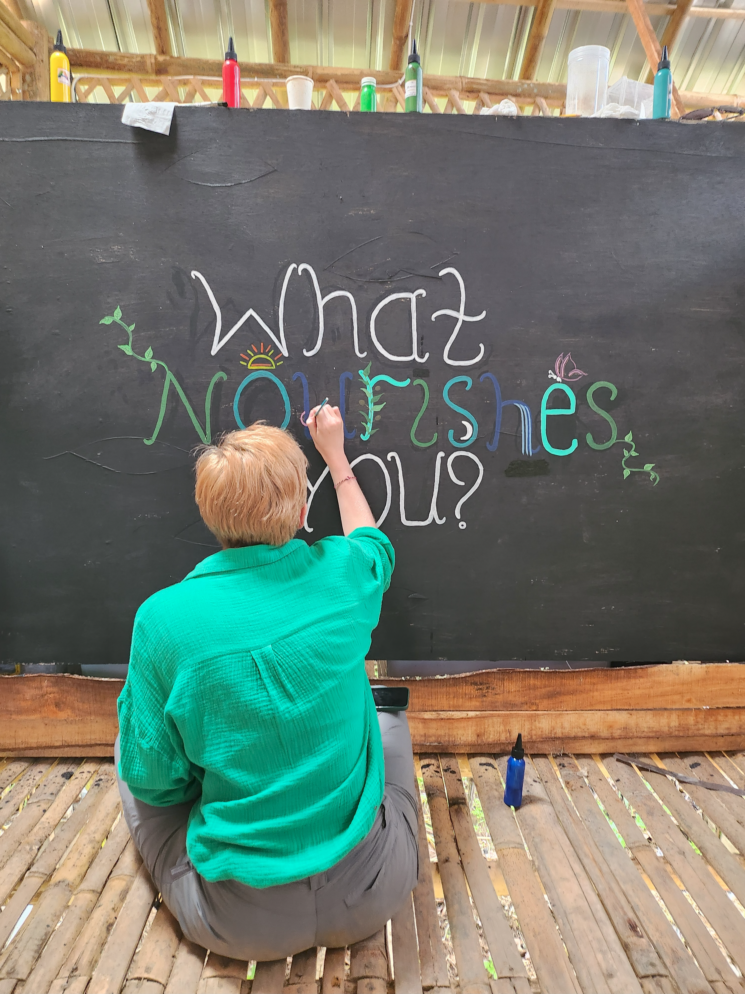 A person with short blonde hair, wearing a turquoise shirt and gray pants, is kneeling on a wooden floor, drawing on a large black chalkboard with colorful markers. The chalkboard has the words 'What Nurtures You?' written on it with decorative eleme
