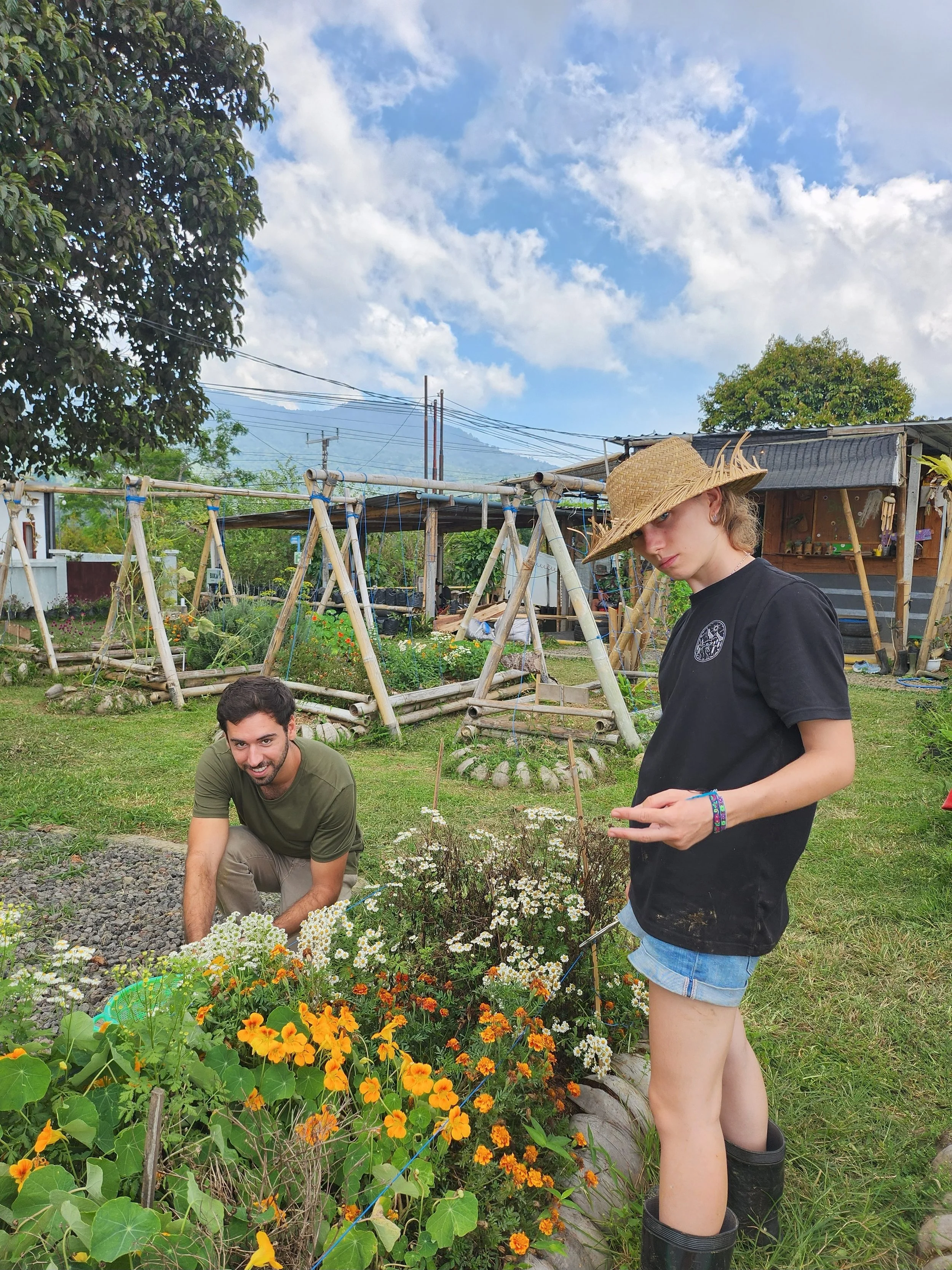 Two people gardening in a backyard with flowers and a wooden swing set. One person is squatting and smiling, the other is standing and wearing a straw hat.