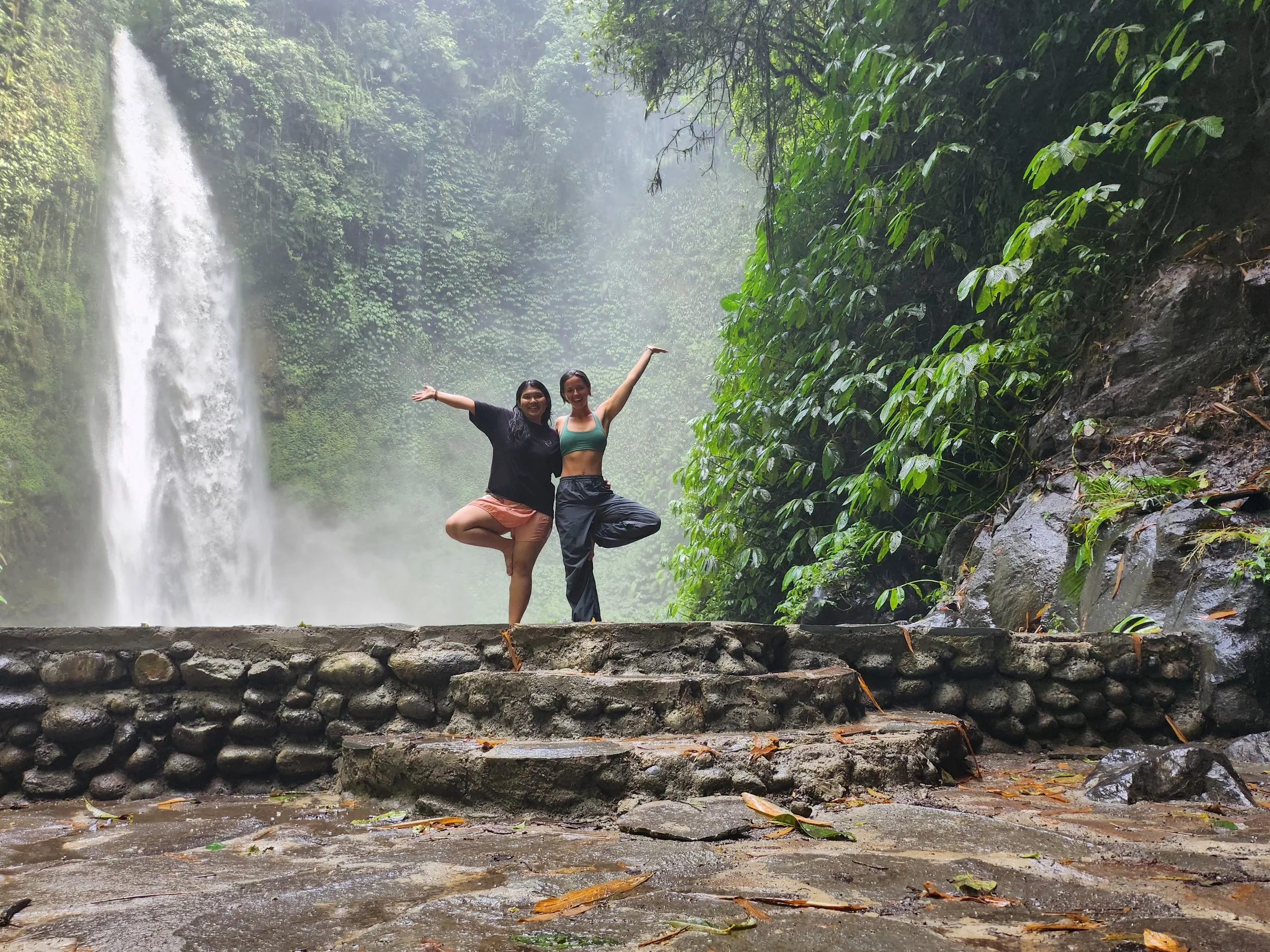 Two women stand on a stone platform with a Nungnung waterfall and lush green jungle in Plaga, Bali, Indonesia in the background. They are posing with one leg raised and arms outstretched, smiling at the camera.