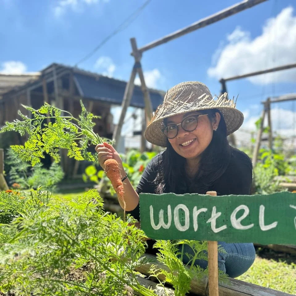 A woman wearing glasses and a straw hat kneeling in a garden, holding a large carrot. There is a green sign with the word 'wortel' in front of her, and various plants and garden structures are visible in the background.