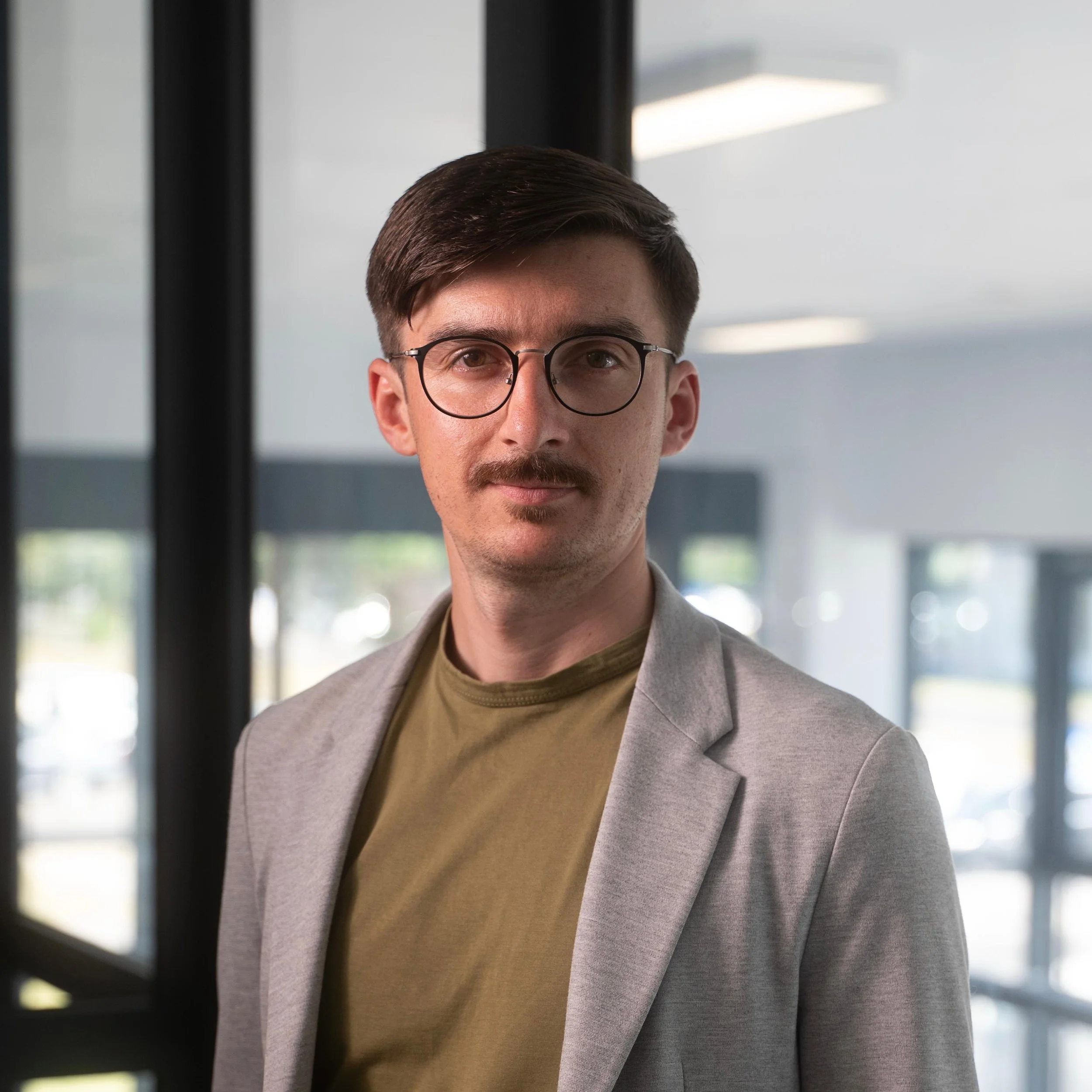 A young man with glasses and a mustache wearing a beige blazer and olive-green shirt, standing indoors near large windows.