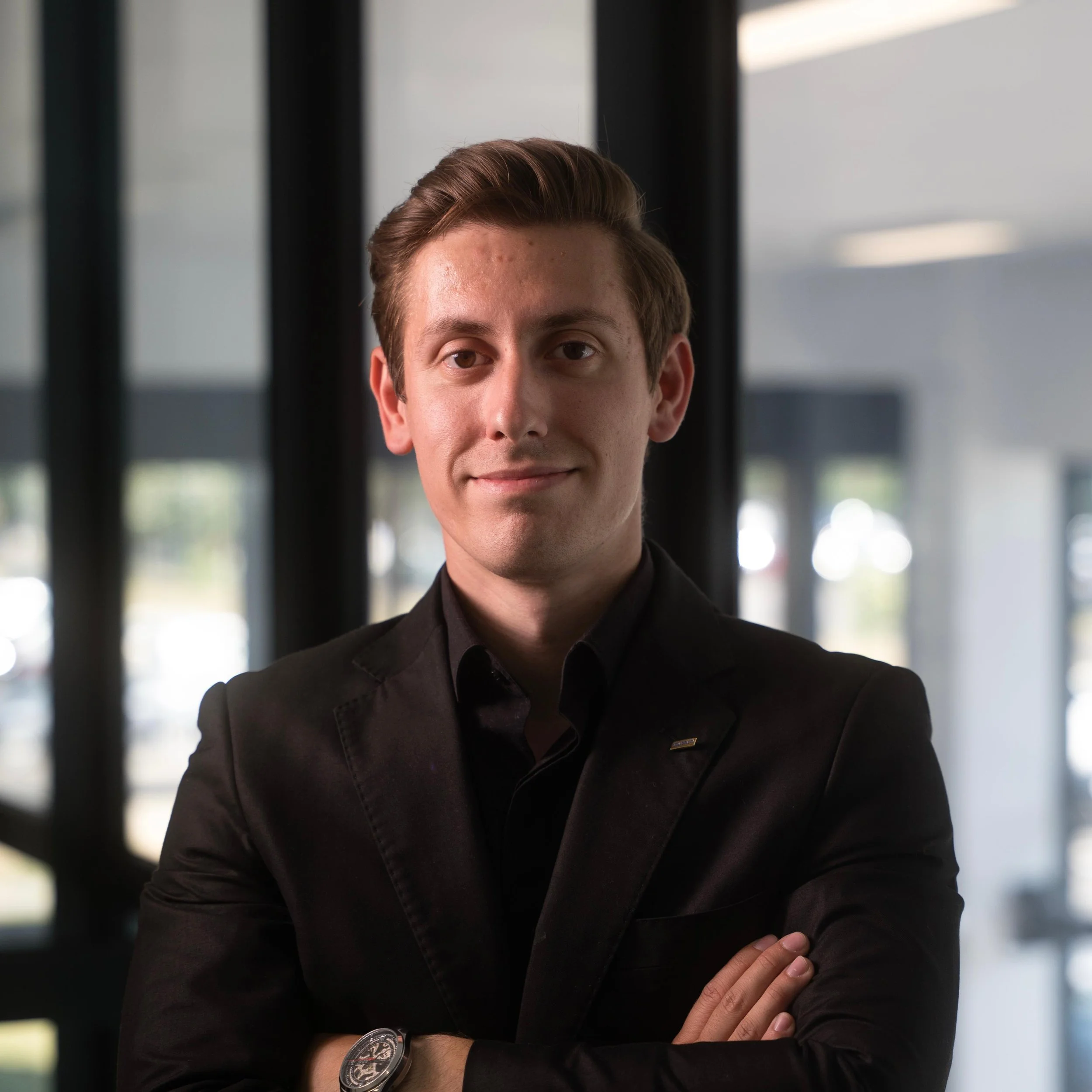 Young man in black suit with arms crossed, standing indoors with blurred windows behind him.