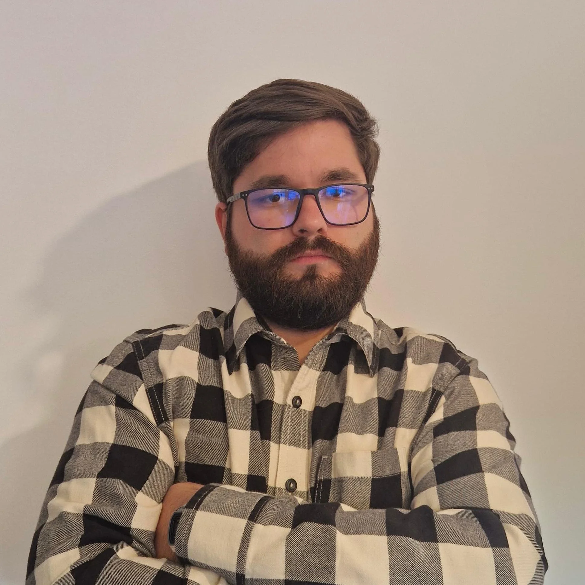 Man wearing glasses and a checkered shirt with arms crossed standing against a plain light-colored wall.