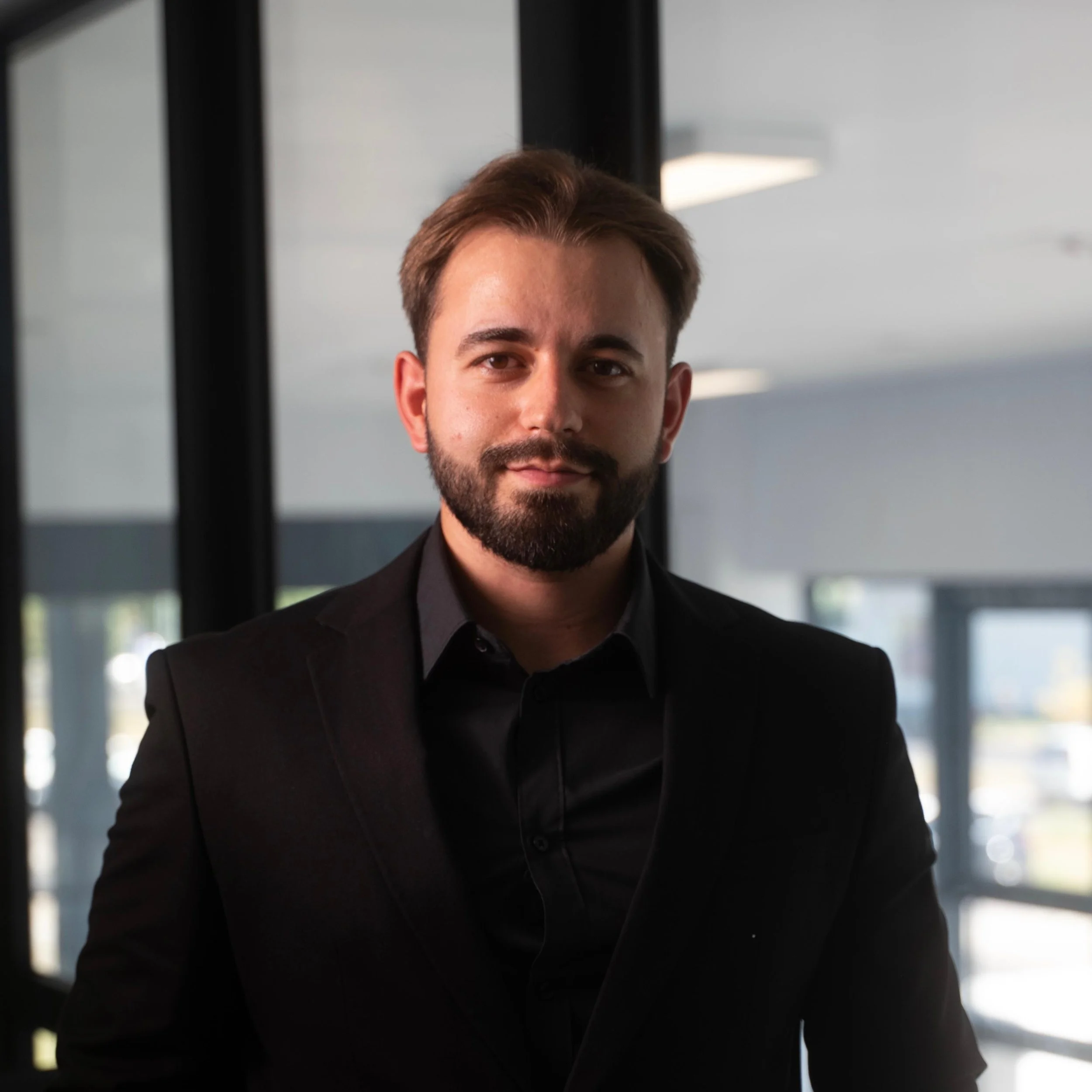 A young man with a beard and short brown hair, wearing a black blazer and black shirt, standing indoors near glass windows.