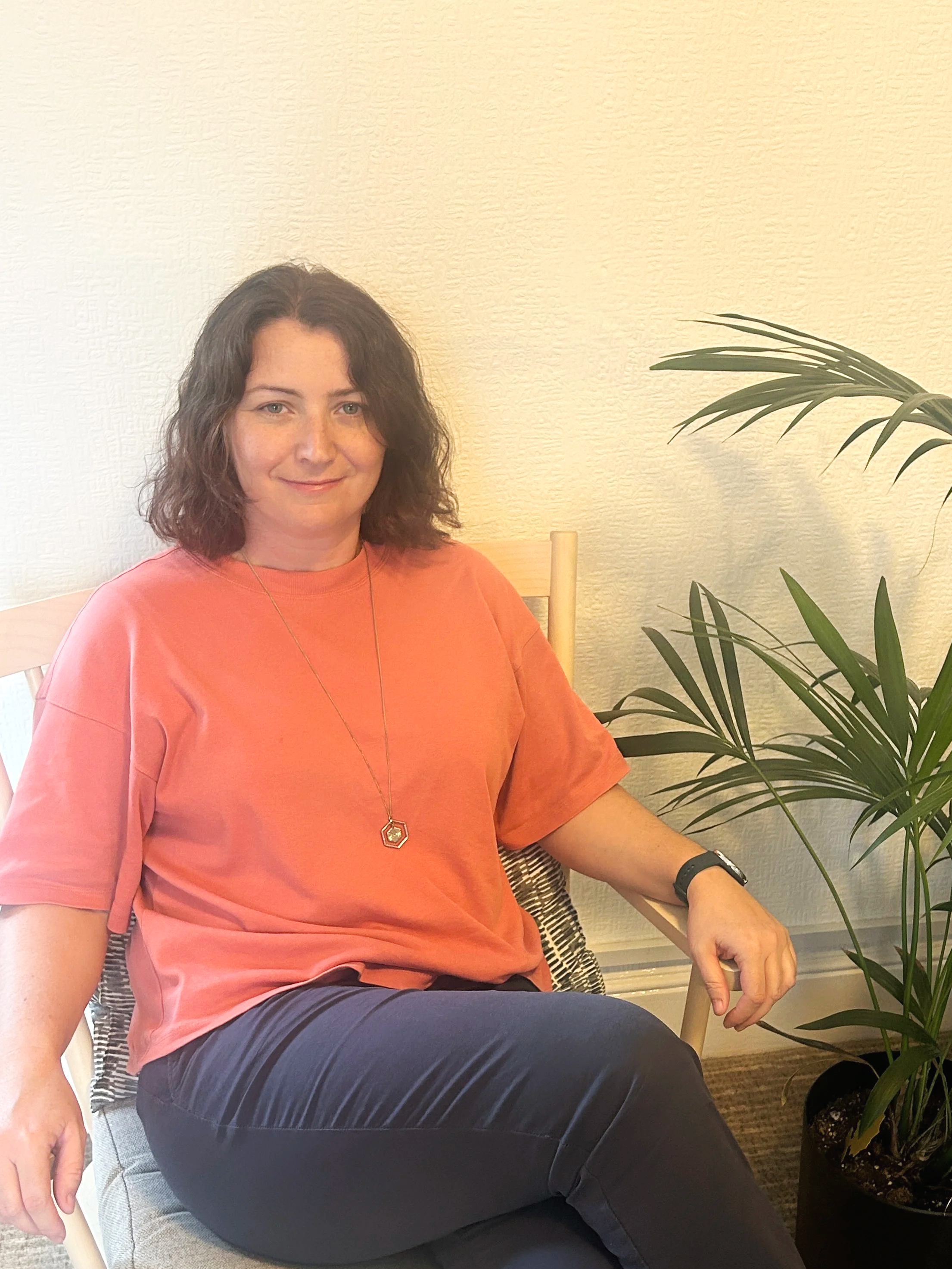 A woman with shoulder-length curly brown hair, wearing an orange T-shirt, blue trousers, and a necklace, sitting on a wooden chair next to a large green potted plant, in a room with beige walls.