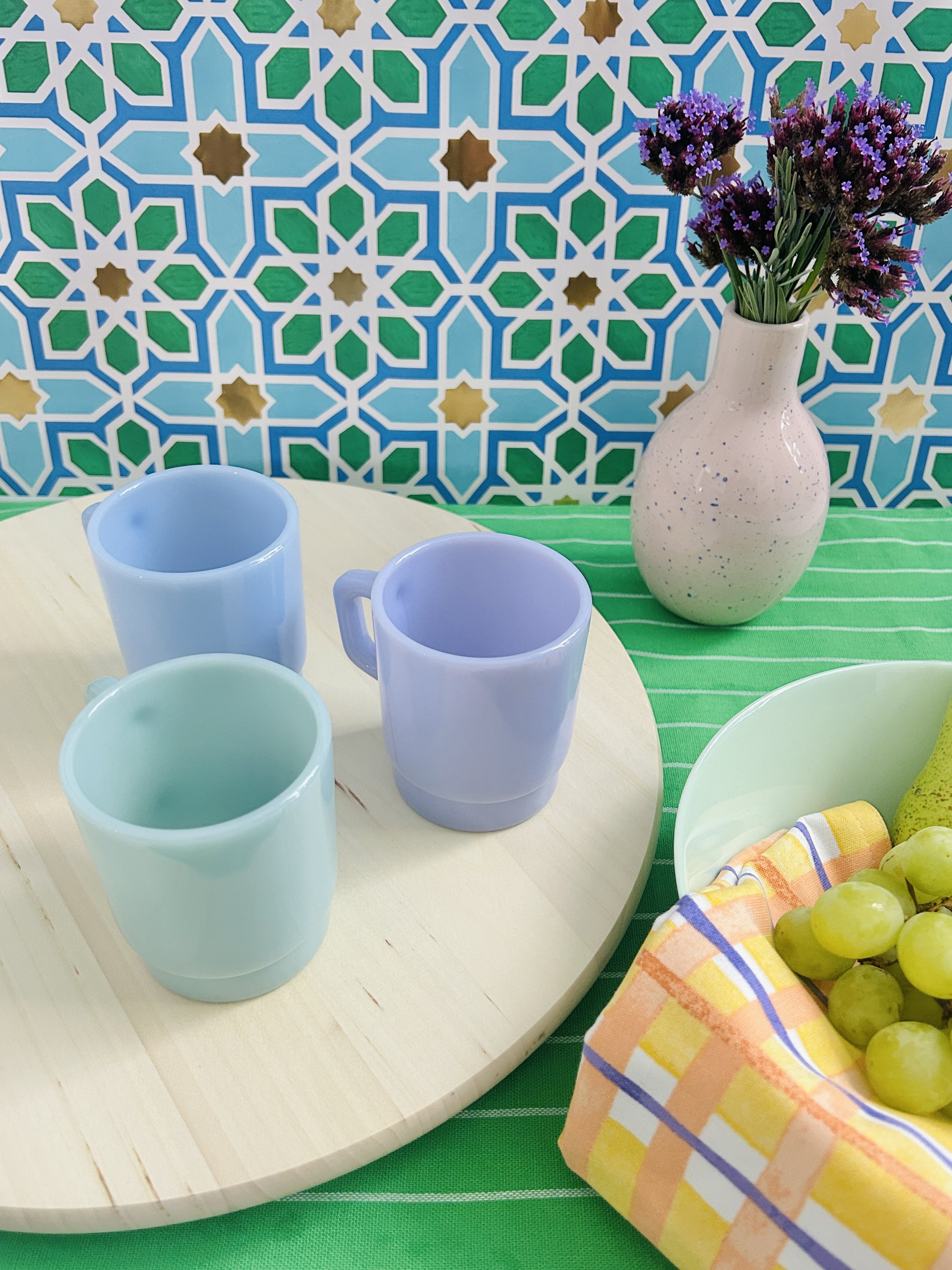 Colourful table setting with pastel-coloured cups, a vase with purple flowers, and a bowl of green grapes against a patterned tile background.