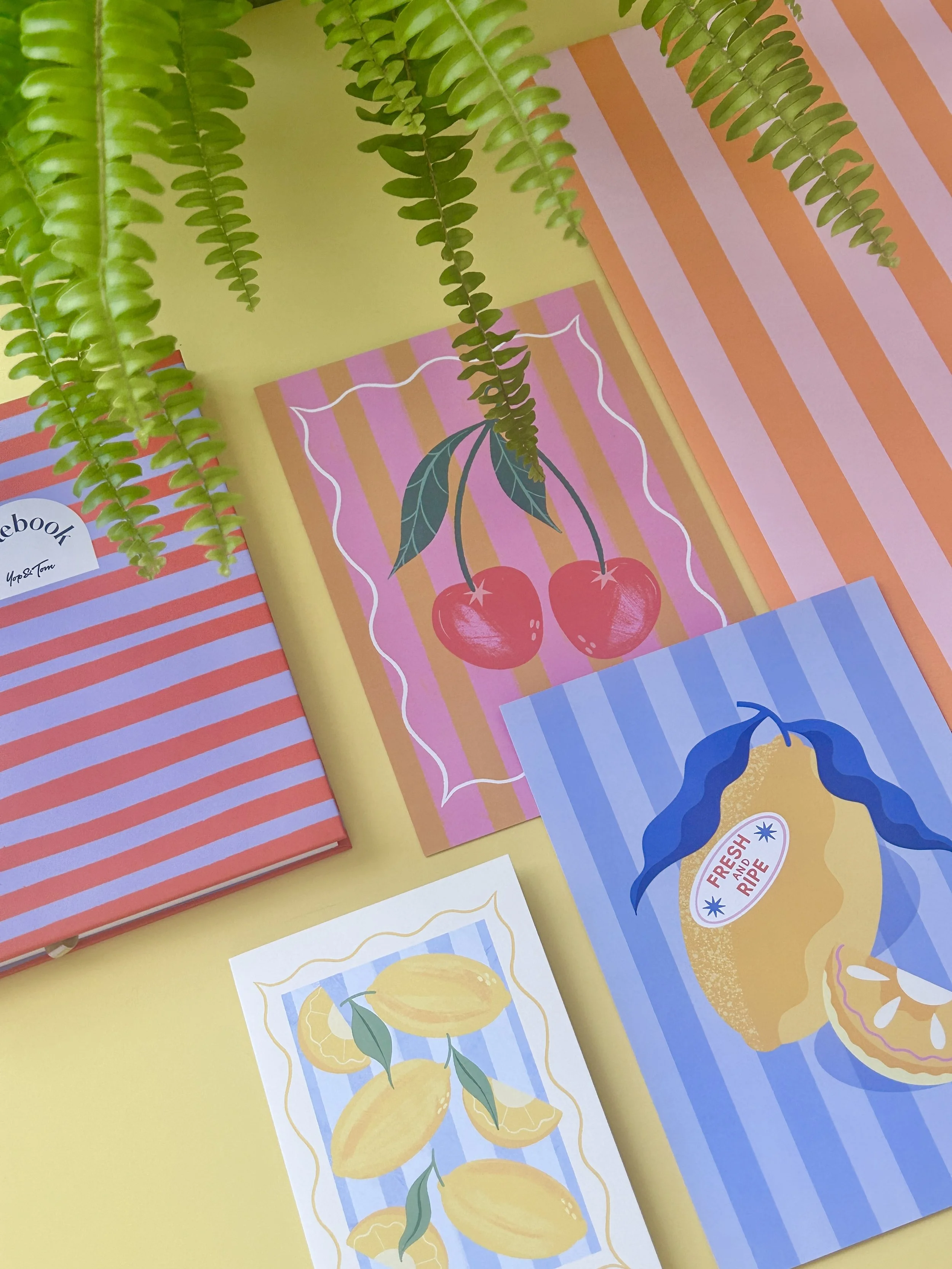 Brightly coloured illustrated fruit-themed stationery and illustrations on a yellow background, including cherries, lemons, and a grapefruit, with green hanging fern leaves above.