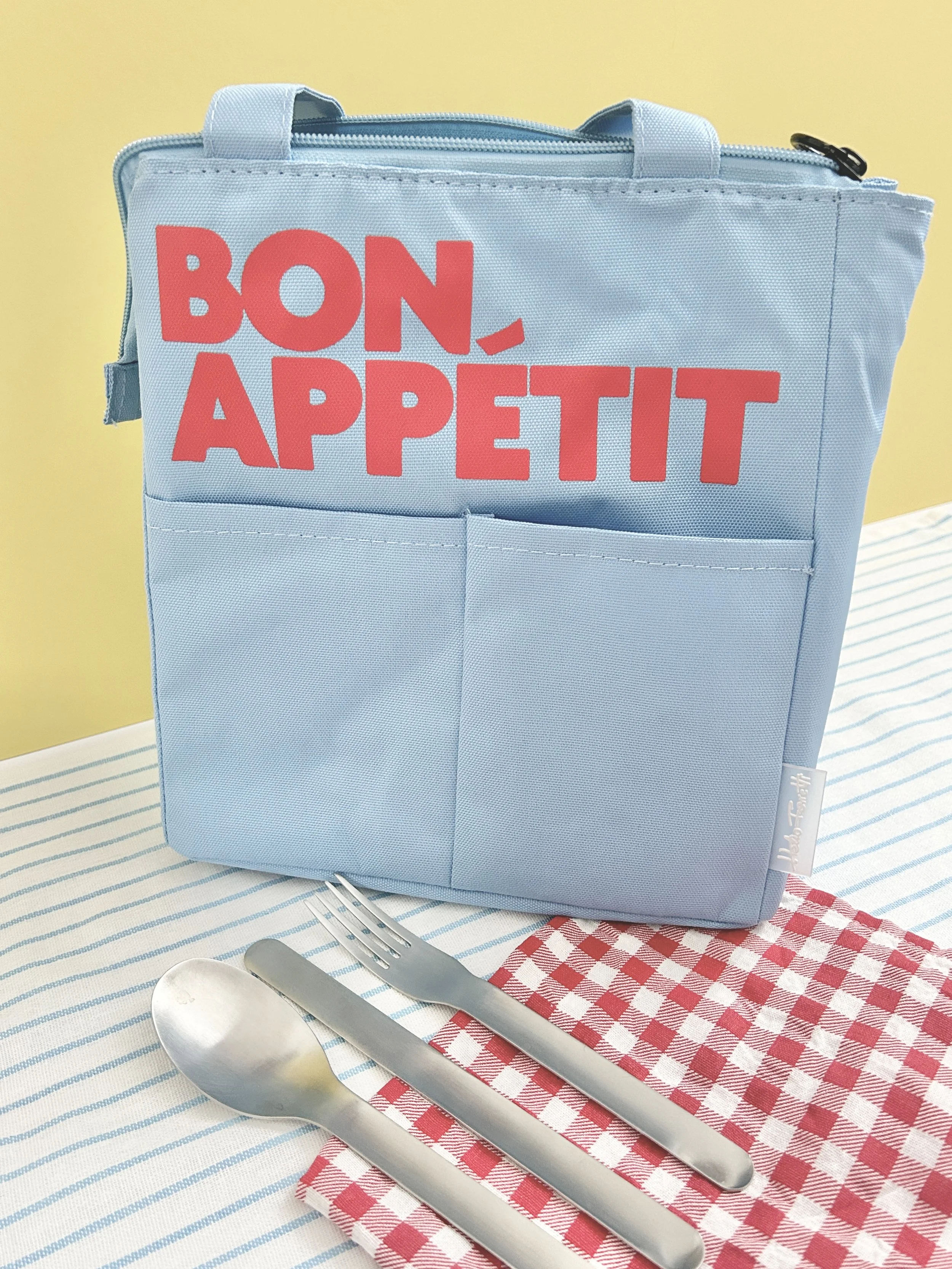 A light blue insulated lunch bag with 'Bon Appétit' written in red letters on the front, placed on a table with a red and white checkered napkin, silver spoon, fork, and knife, with a yellow wall in the background.