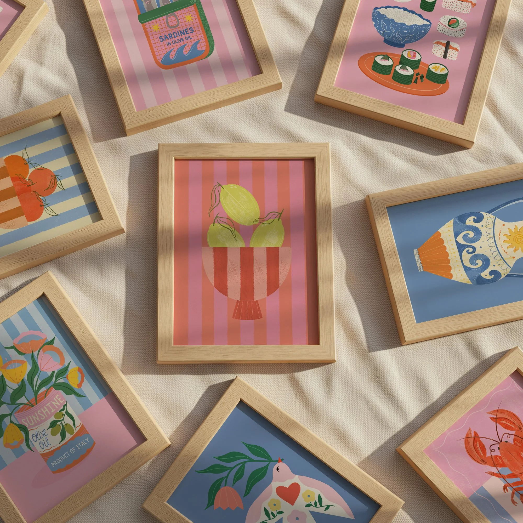 Collection of framed colourful illustrations including a jar of sardines, sushi rolls, tomatoes, a vase with fruit, a potted plant, a ceramic container, a flowering plant, and a lobster, all displayed on a fabric surface.