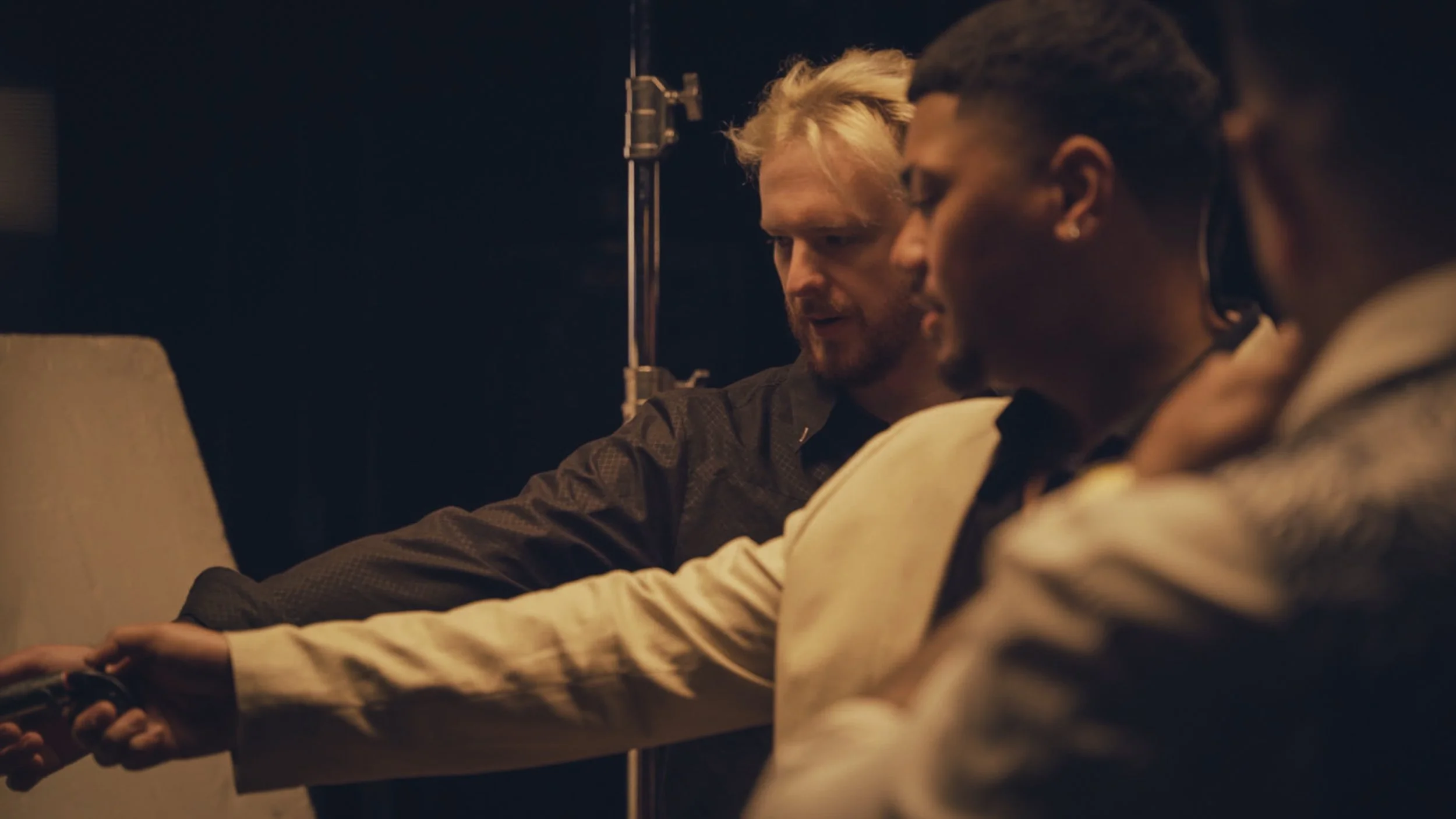Three men are stood together, leaning forward, and holding a prop, appearing focused while about to shoot a scene in a dark room.
