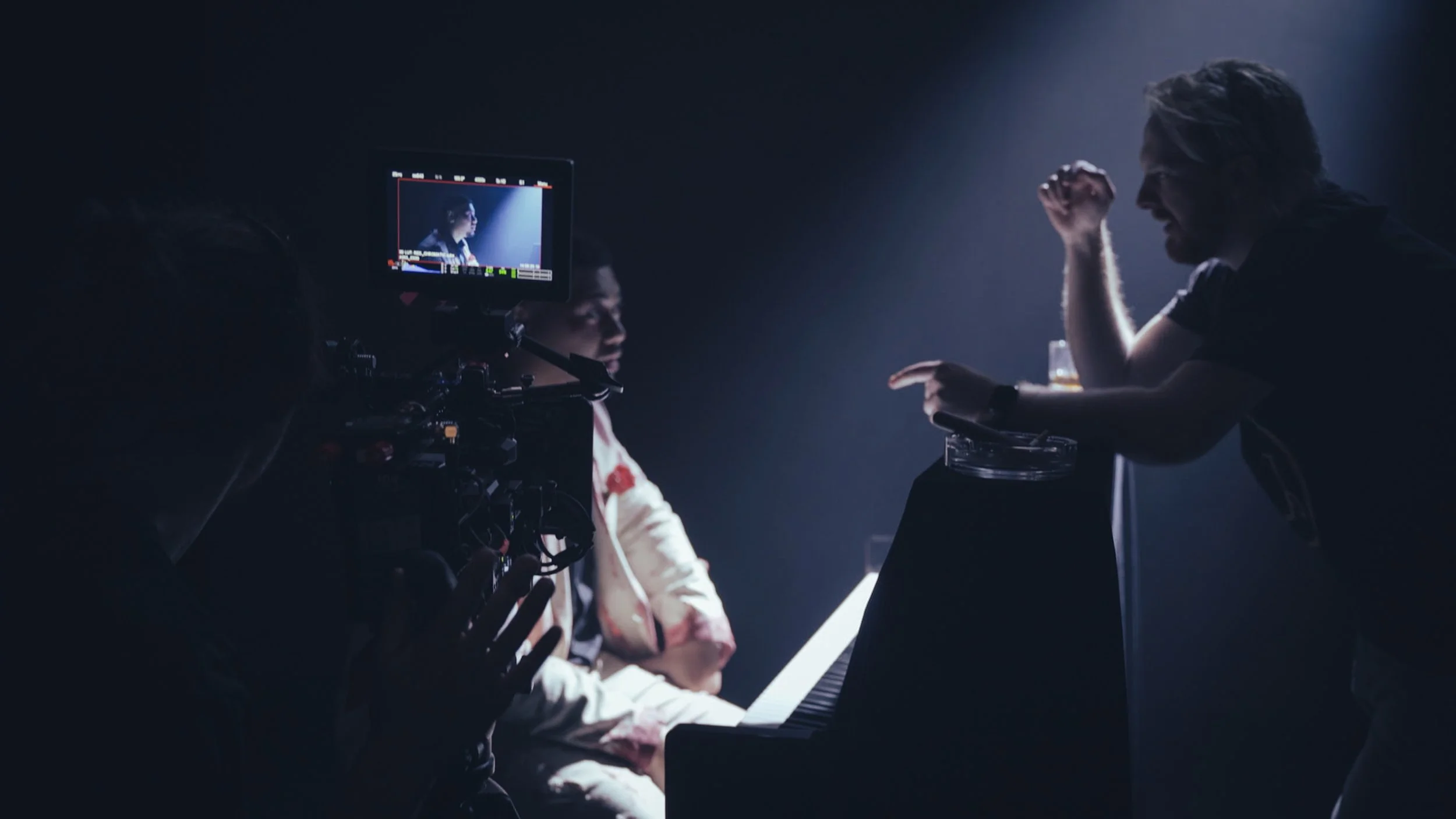 Behind-the-scenes shot of a film or TV set with a man playing piano, a director speaking to him, and a crew member operating a camera in a dimly lit studio.