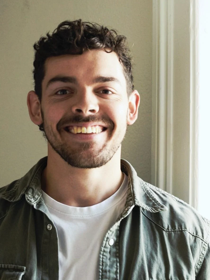A young man with short curly hair and a beard, smiling, wearing a beige shirt over a white t-shirt, standing near a window.