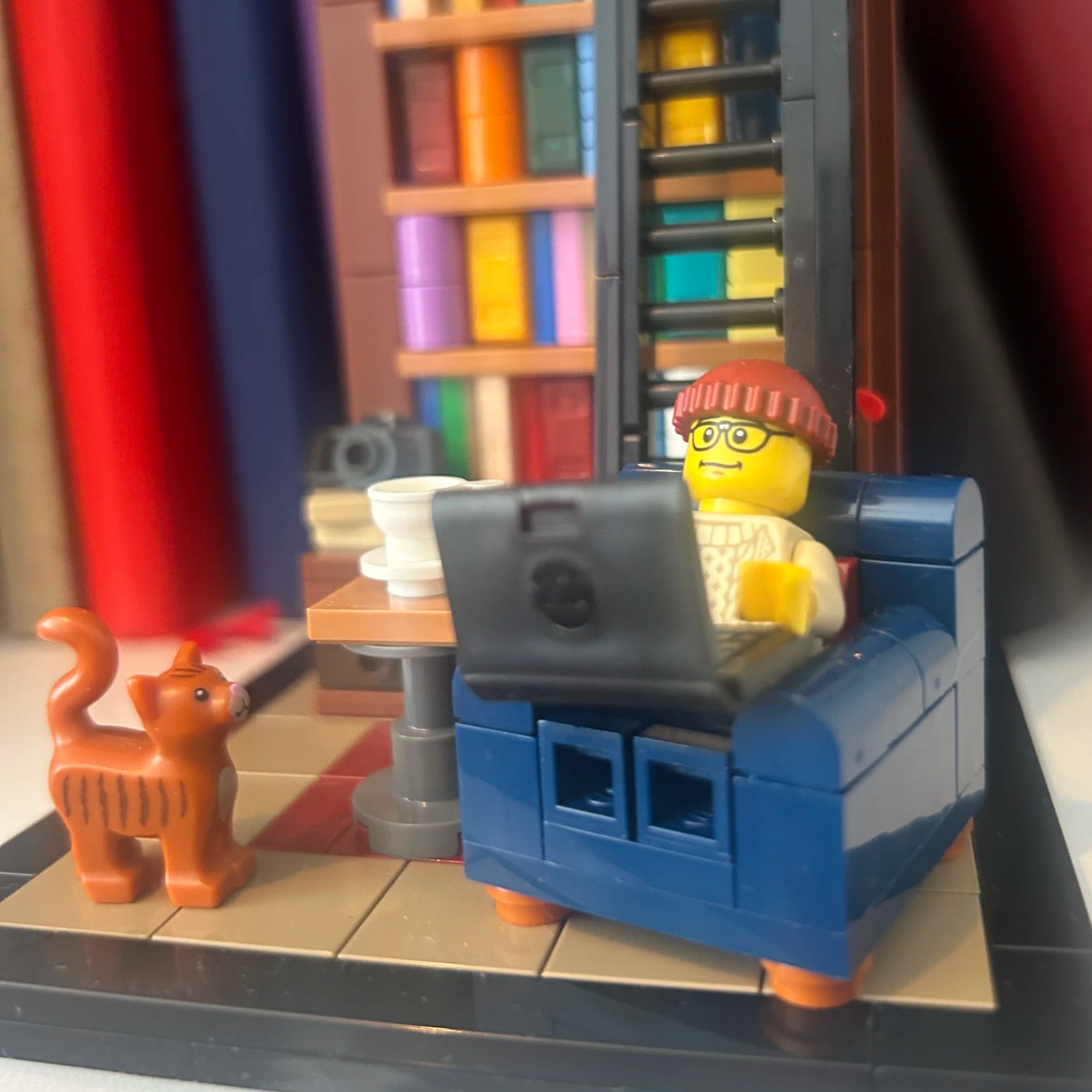 A lego minifigure resembling Stephen sits on a blue lego chair reading a laptop with a ginger lego cat at his feet