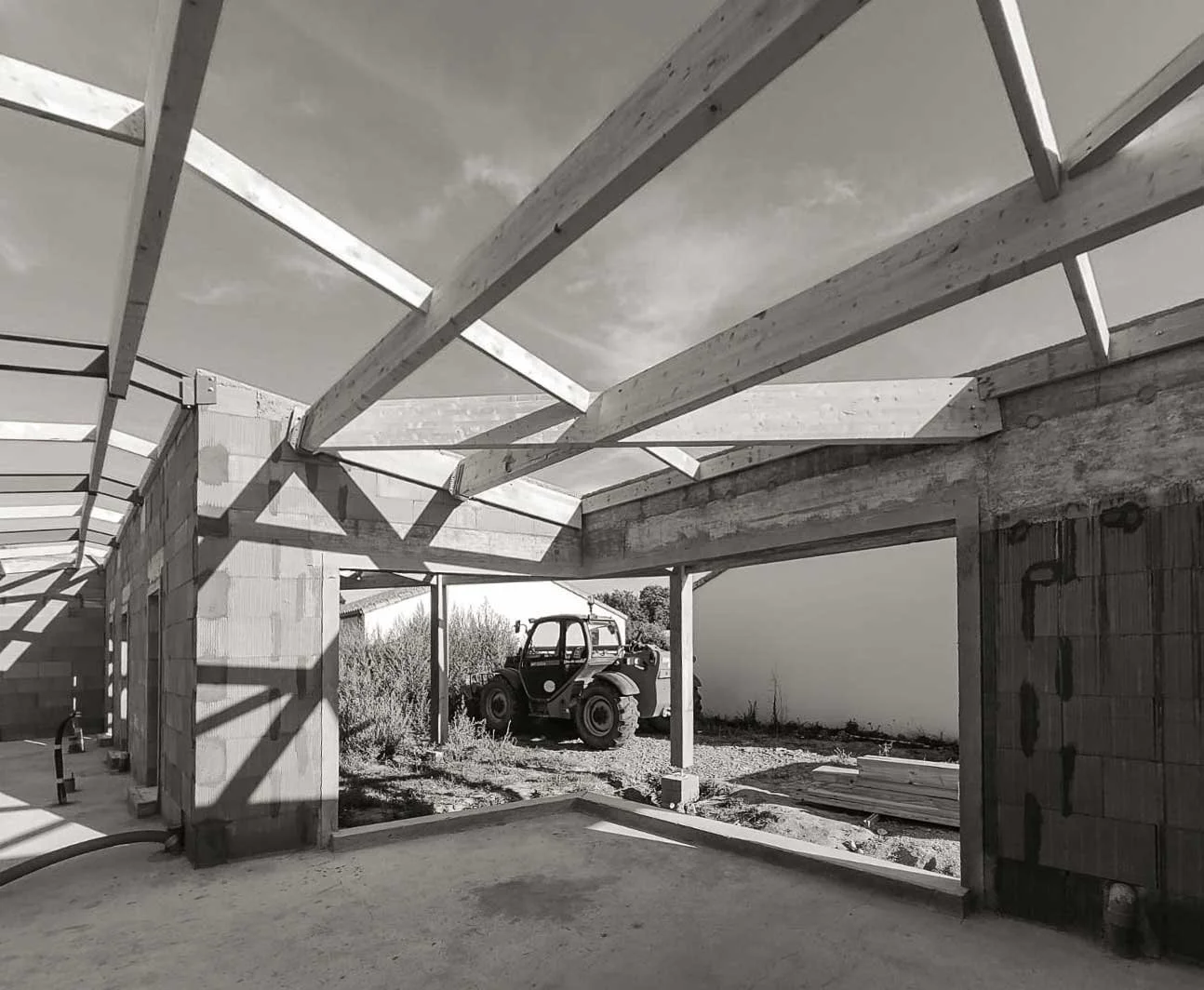 Construction d'une structure en bois avec un bâtiment en béton en arrière-plan, un tracteur à l'extérieur, sous un ciel nuageux.