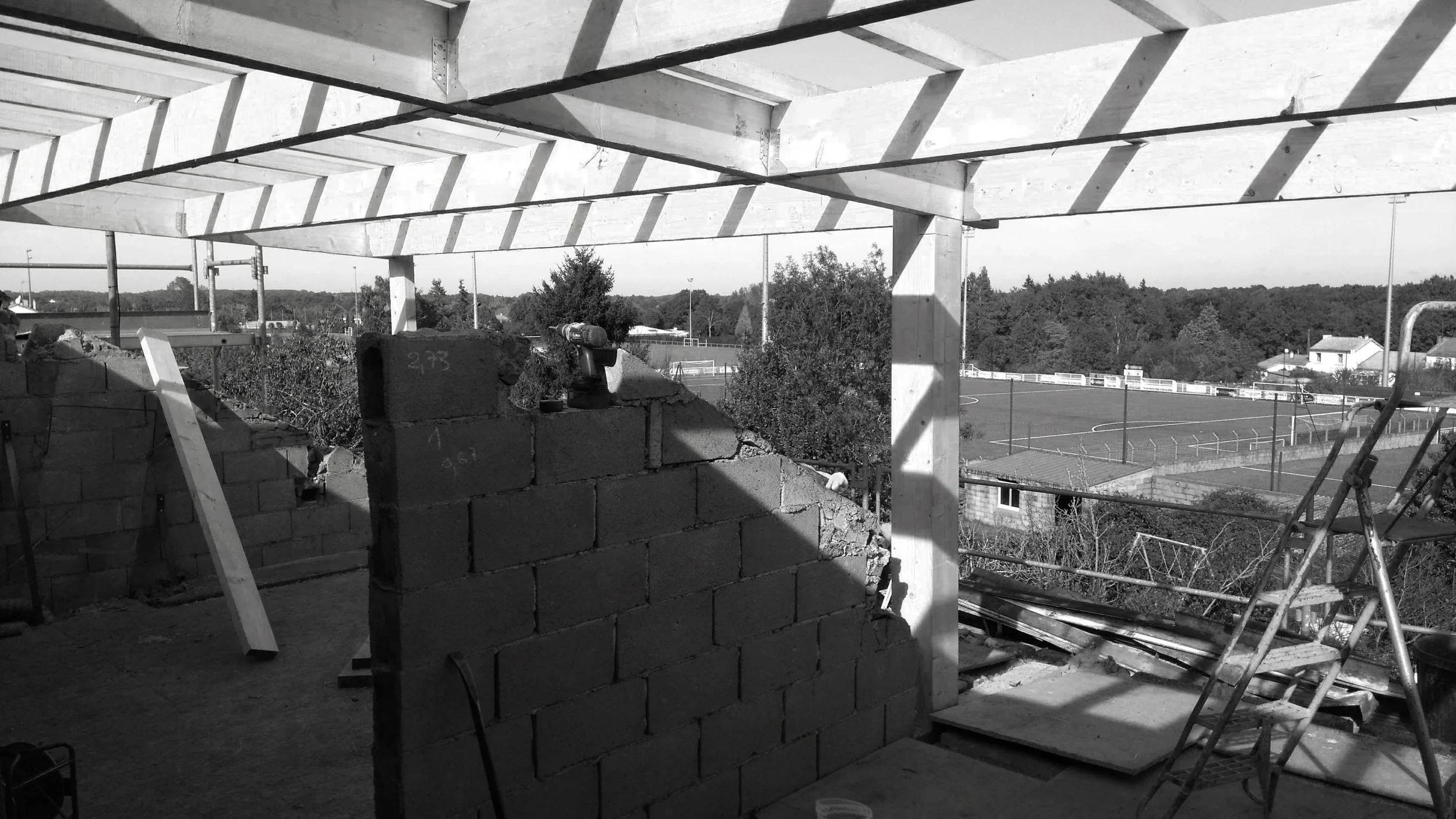 Construction en cours sur un bâtiment, vue sur un terrain de sport avec terrains de tennis et de football en arrière-plan, sous un ciel dégagé.