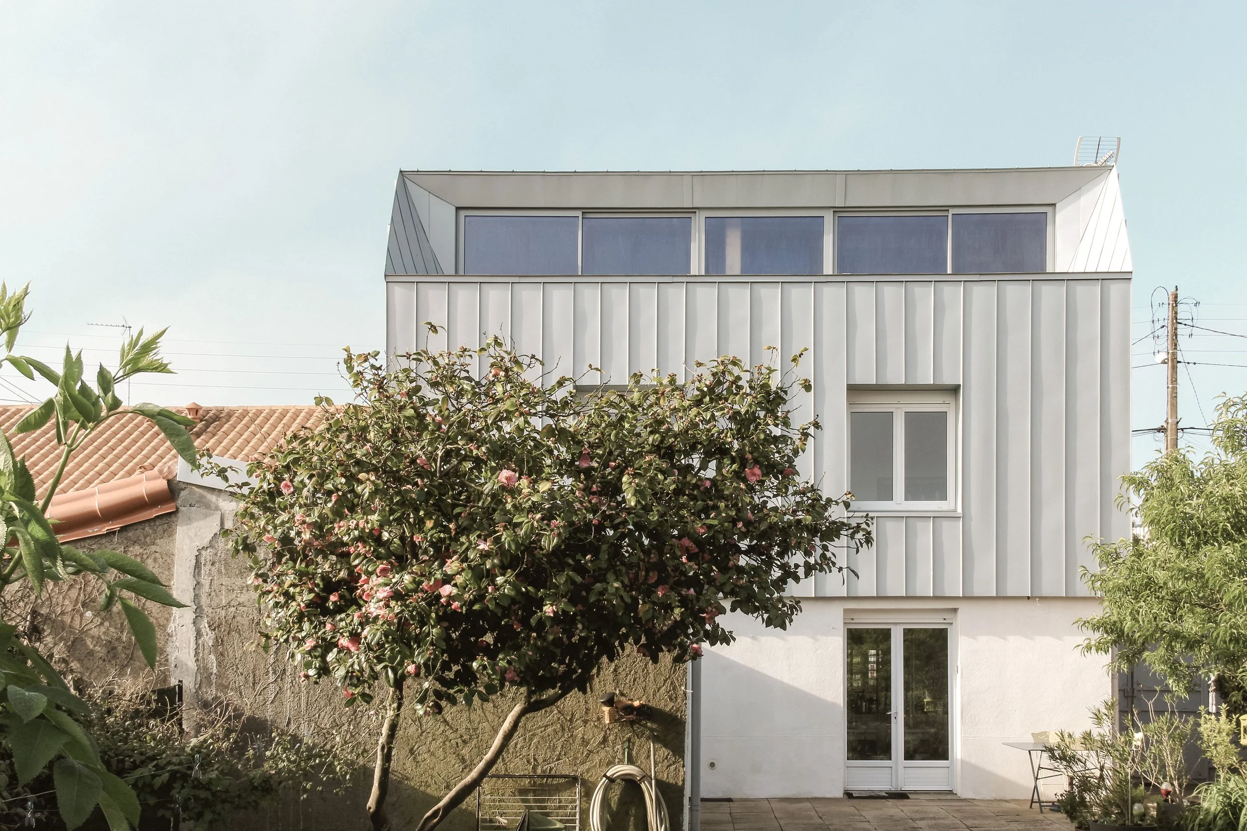 Vue d'une maison moderne avec une façade blanche, une grande baie vitrée et un toit en métal, entourée d'arbres et d'un jardin avec un fauteuil et une petite table.