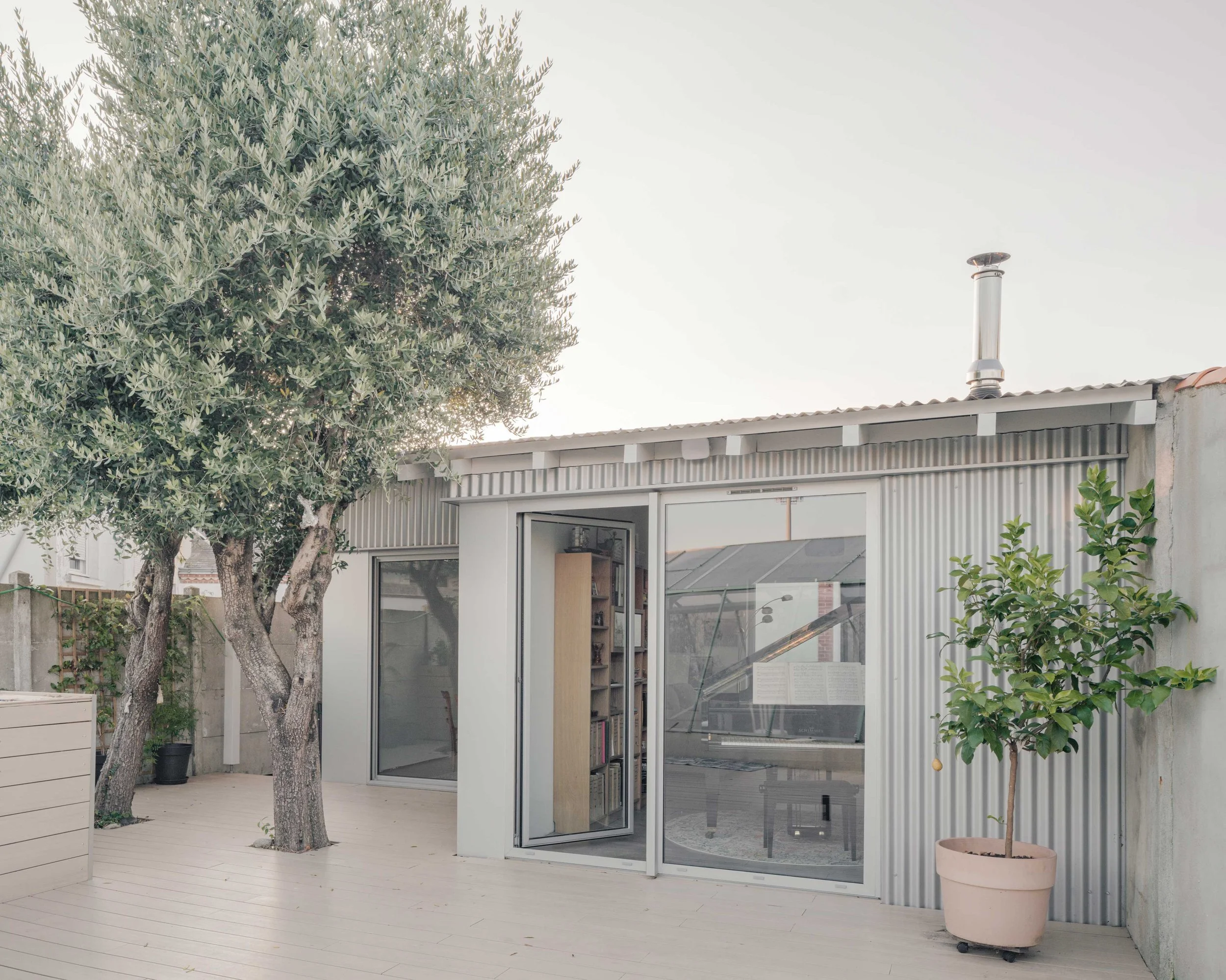 Le patio de derrière de la maison en métal. Deux grands arbres, un en pot et un autre directement dans le sol, entourent la maison. La maison a des murs en métal gris avec de grandes fenêtres en verre. La porte d'entrée est en verre, ouverte, laissan