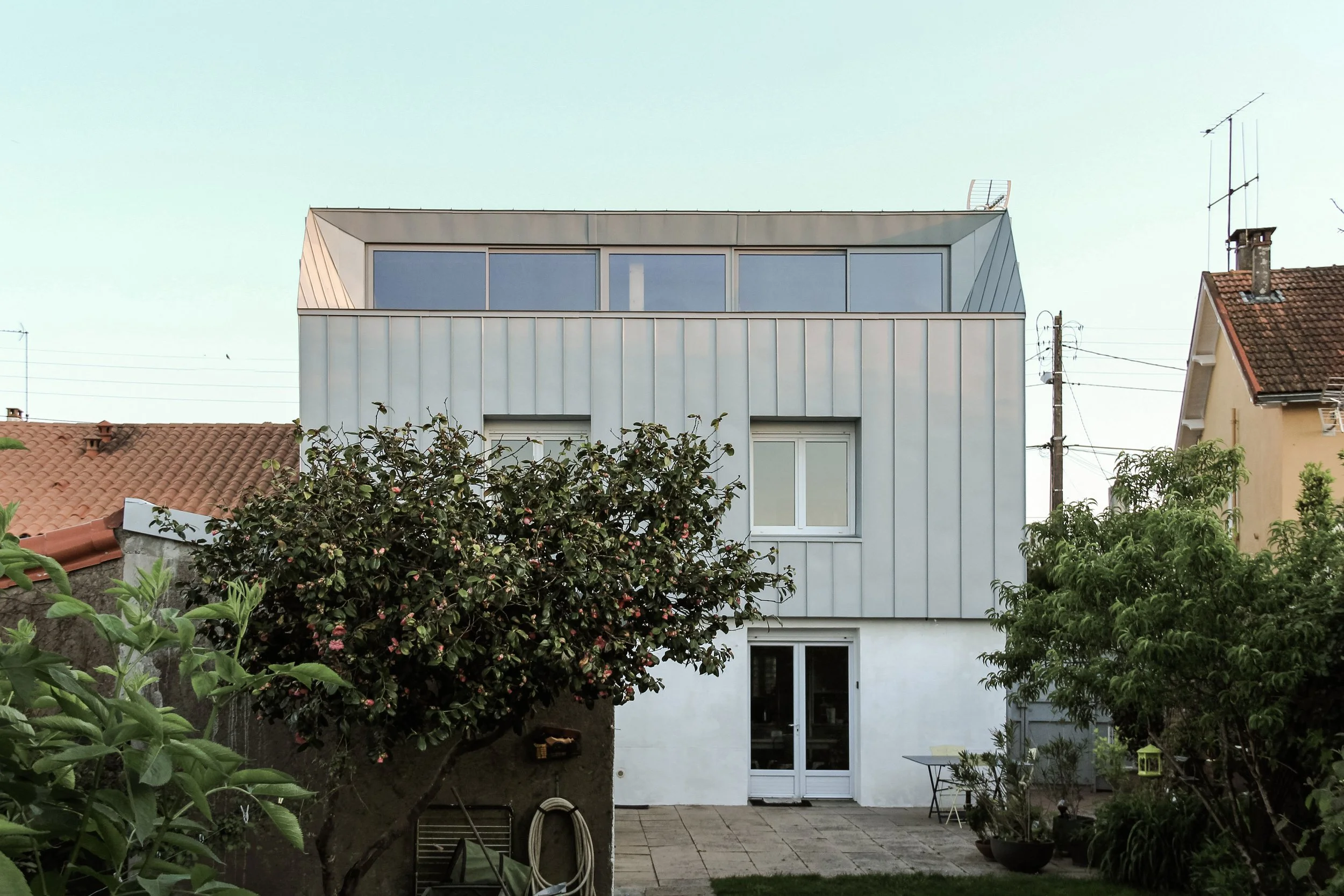 Une maison moderne à plusieurs étages avec de grandes fenêtres, entourée d'un jardin avec des arbres et des meubles de jardin.