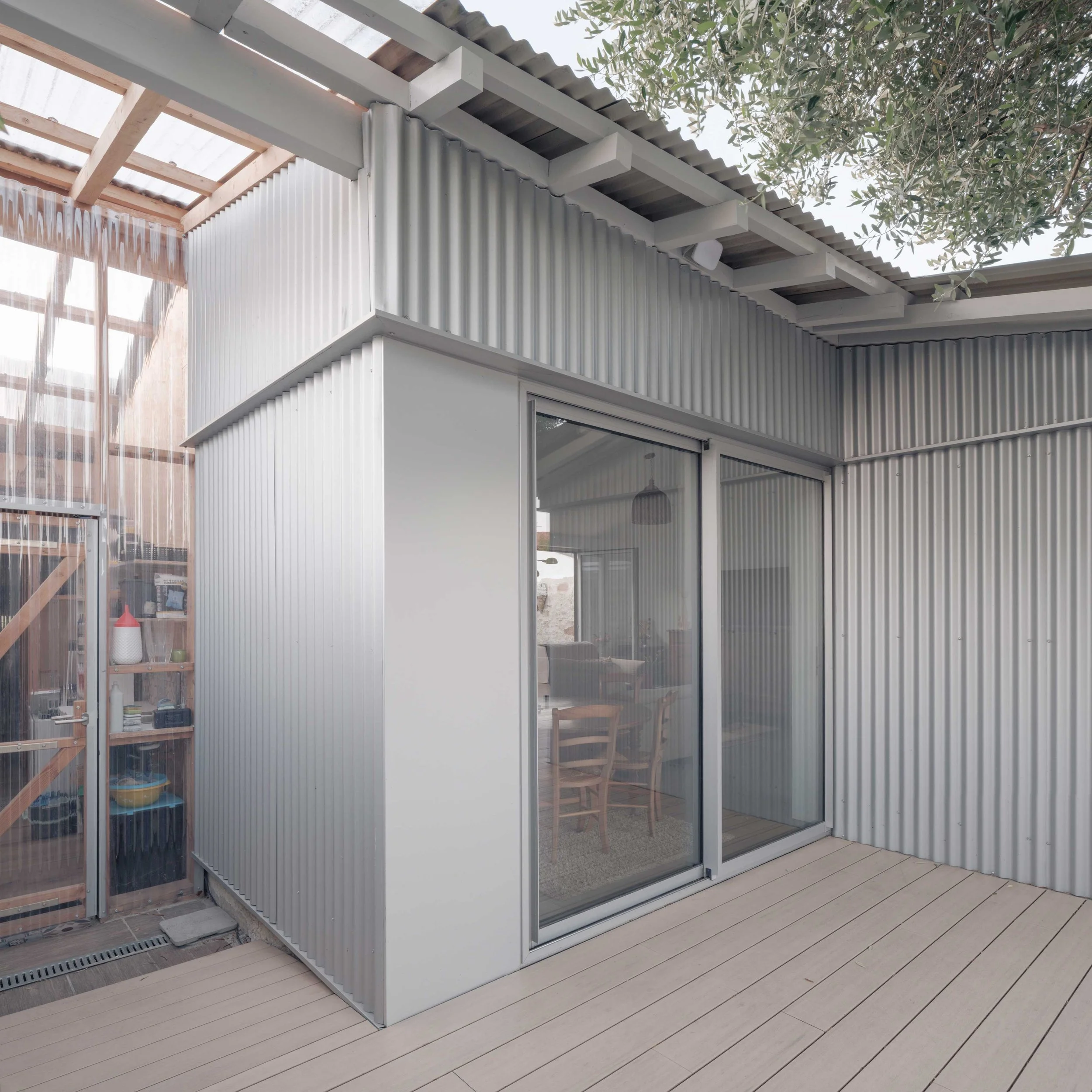 Une structure extérieure en métal avec des murs en panneaux métalliques ondulés, une fenêtre en verre, un patio en bois avec une chaise en bois. La scène est partiellement ombragée par des branches d'arbre